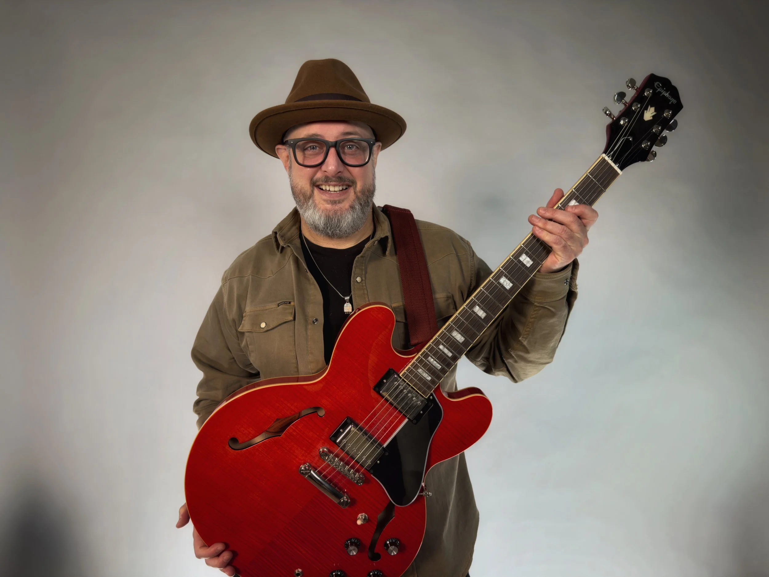 A man with glasses and a beard wearing a brown hat smiles and holds a red electric guitar.