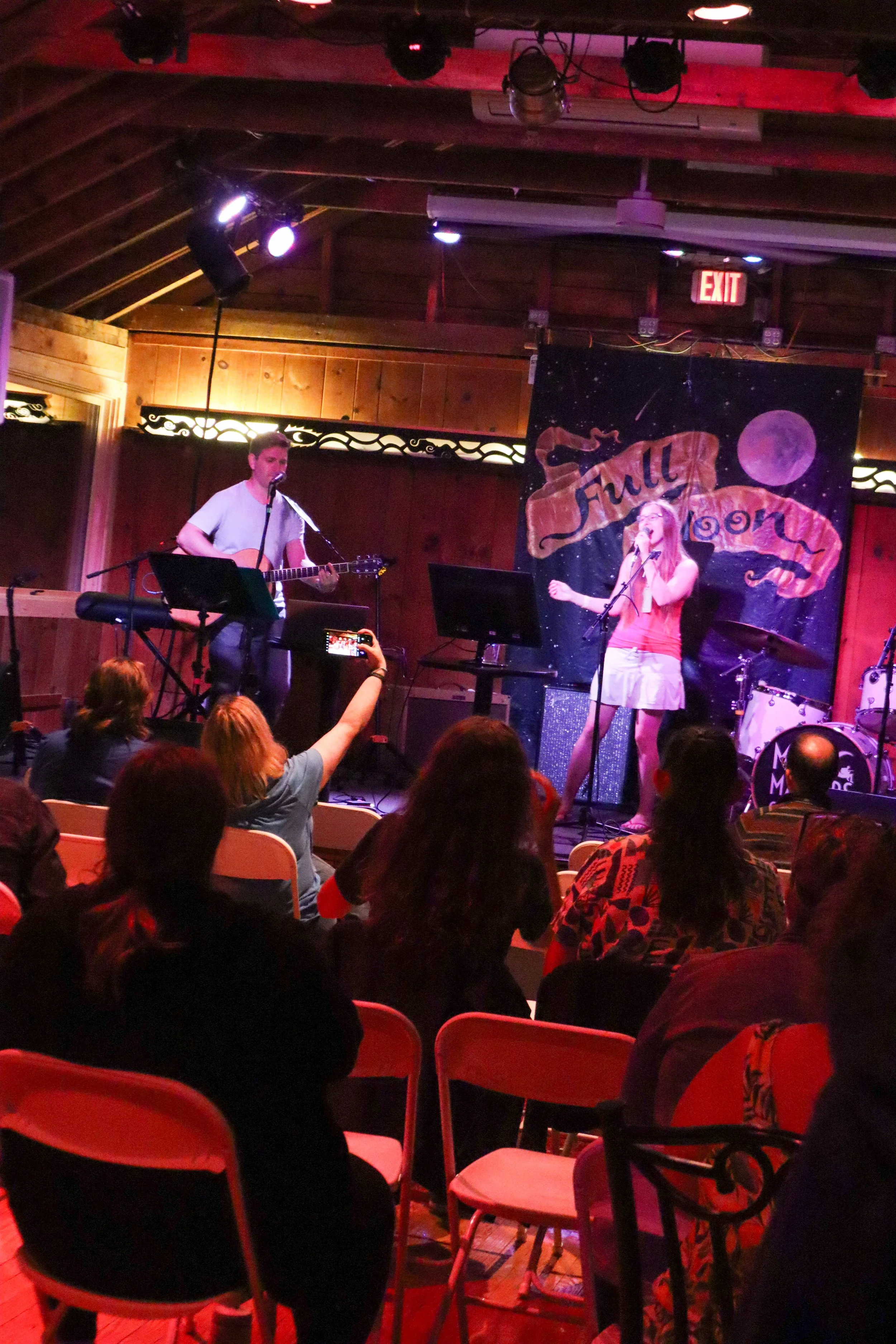 A band performs on stage at a venue with wooden walls and a painted backdrop that says 'Full Moon.' The band includes a female singer and a male guitarist. Audience members are seated and watching, with one person recording the performance on a phone.