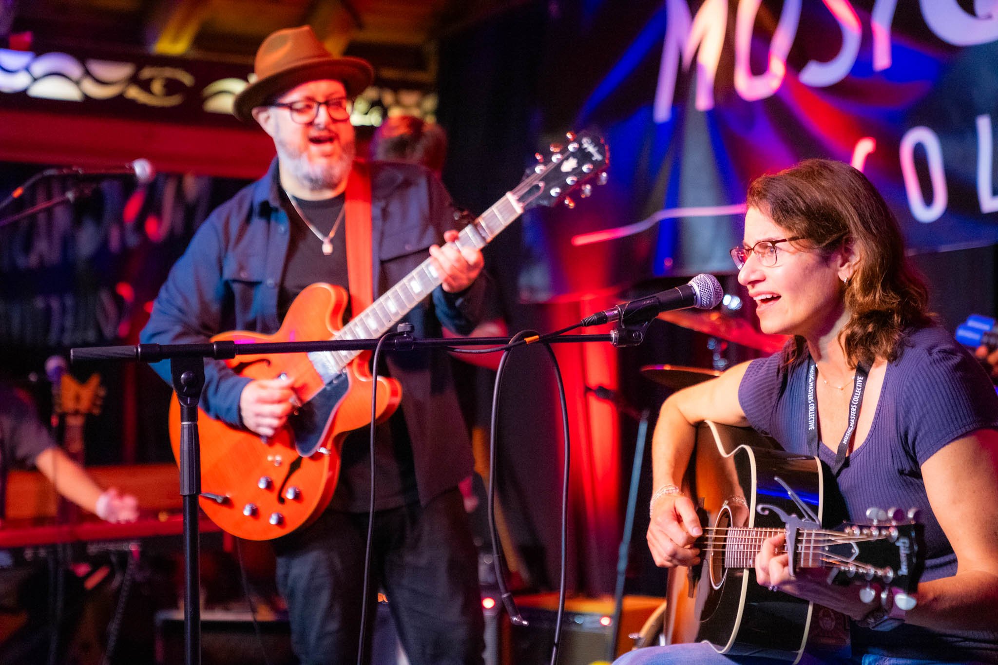 Two musicians performing on stage at a music venue, one man playing an orange electric guitar and another woman singing into a microphone while playing an acoustic guitar, with colorful stage lighting and a neon sign in the background.