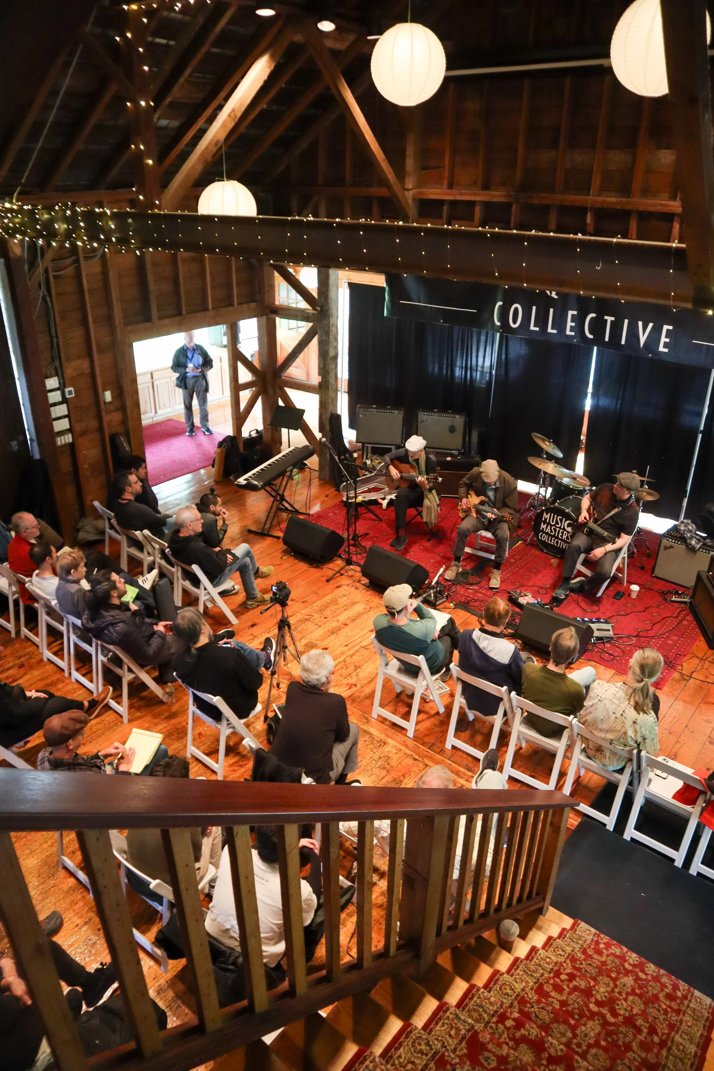 A musical performance takes place in a rustic, wooden indoor venue with an audience seated on white chairs. The band features musicians playing guitars, with drums and amplifiers on stage. The venue is decorated with string lights and paper lanterns hanging from the ceiling.