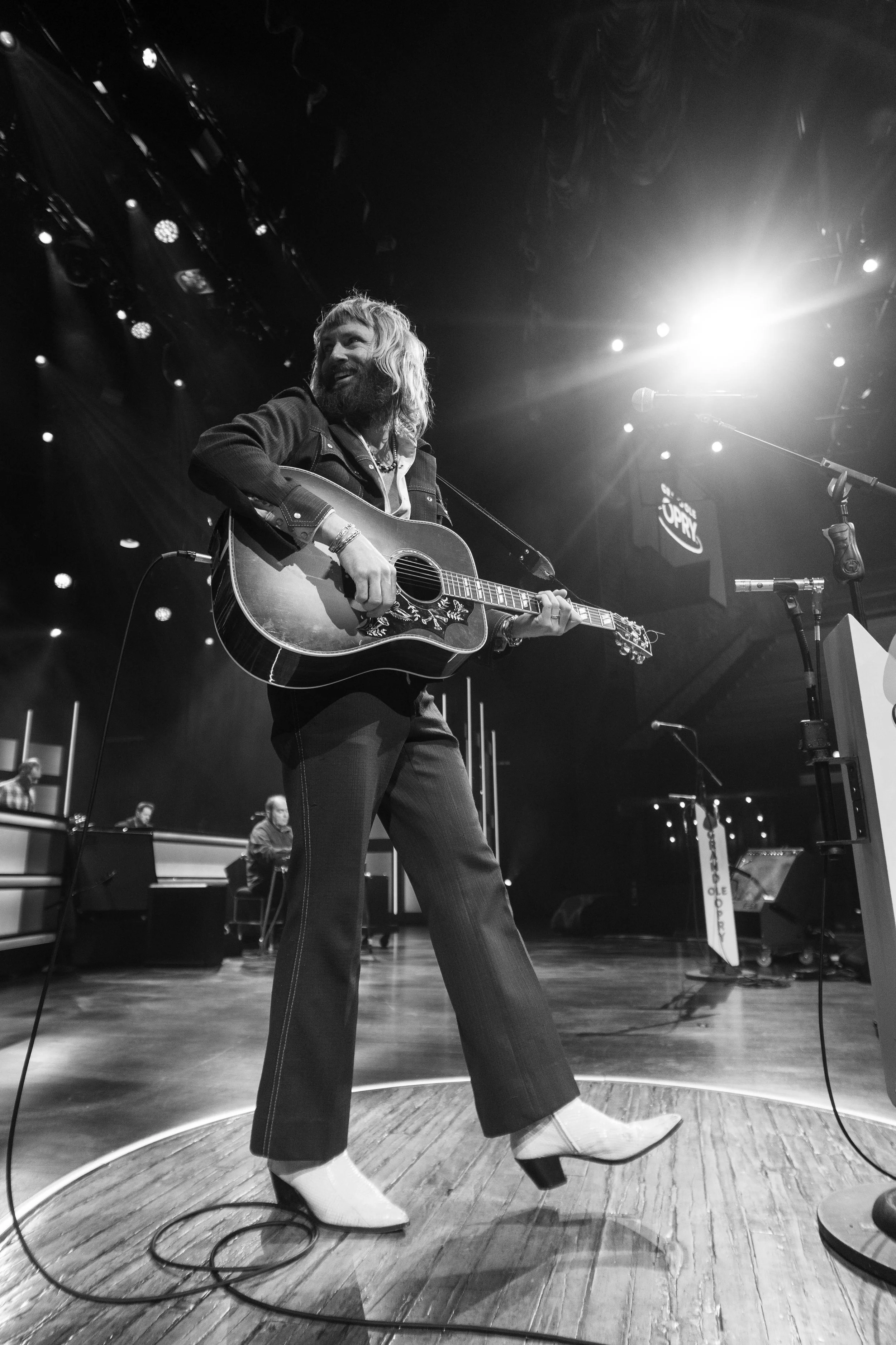 A person with long hair and a beard playing an acoustic guitar on stage during a performance, with stage lights and other musicians in the background.