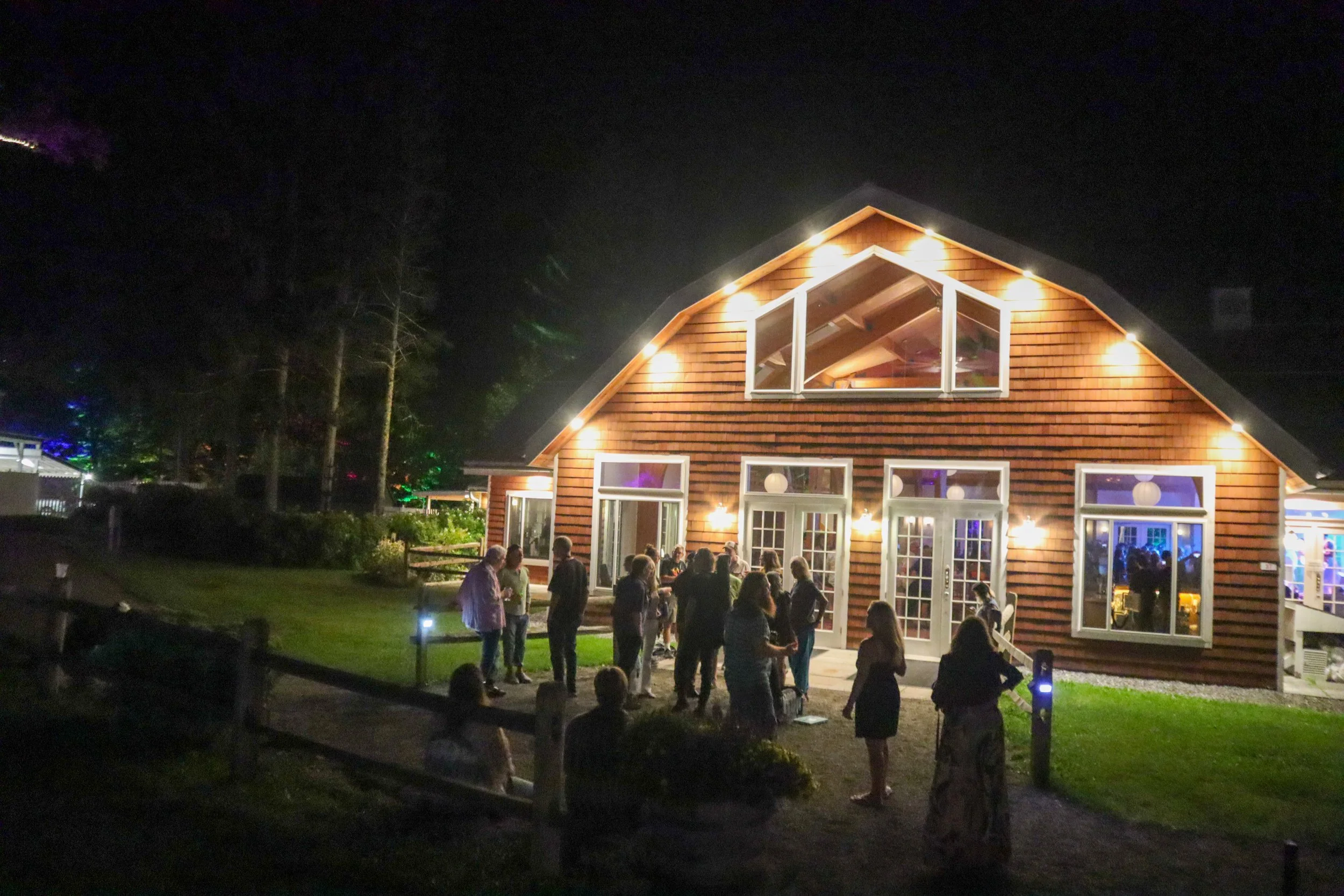 Nighttime outdoor gathering outside a large wooden house with large windows and bright lights, with people socializing on the lawn.