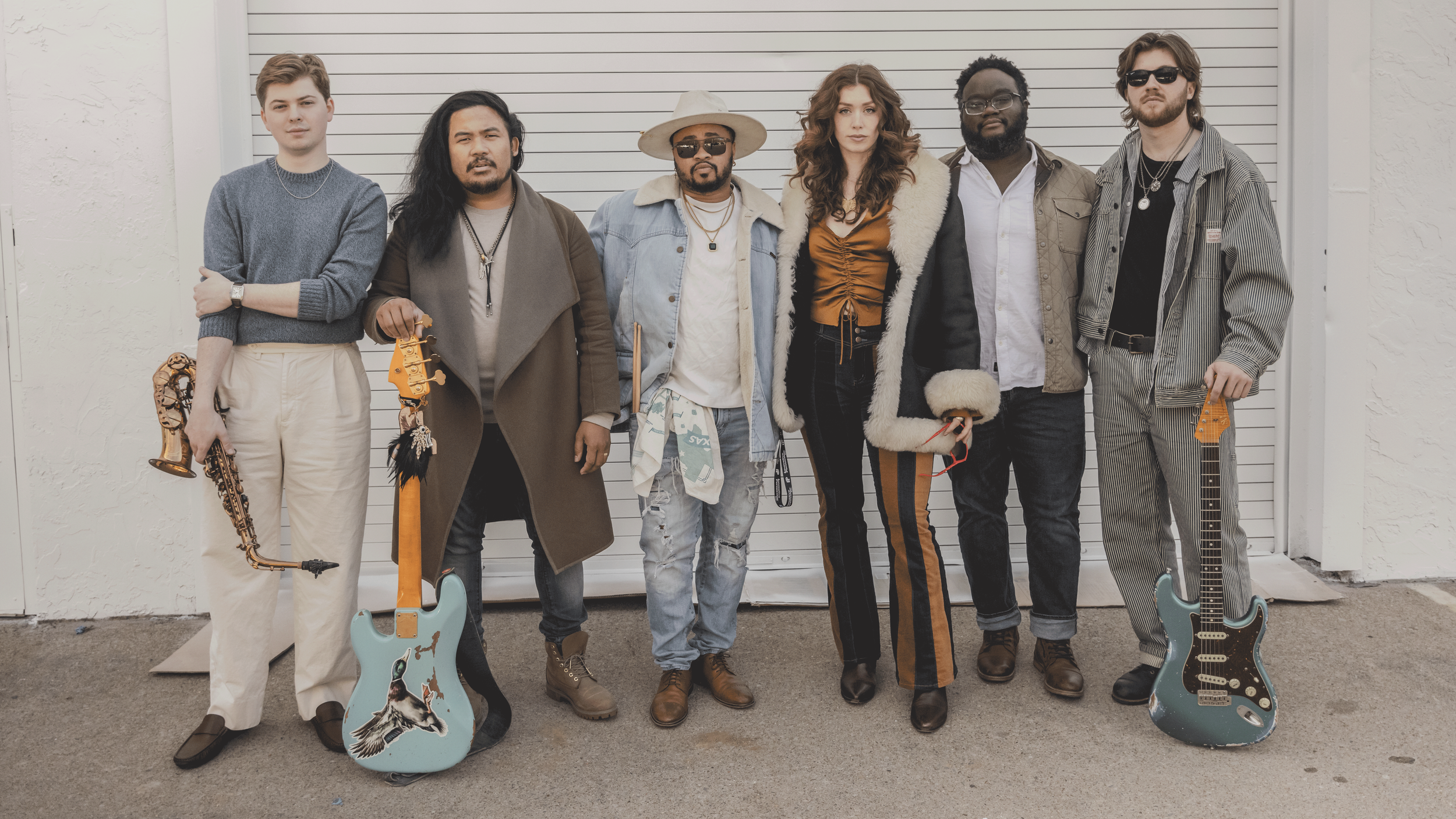 Seven musicians standing in front of a white roll-up door, holding guitars and musical instruments, dressed in casual and stylish clothing.
