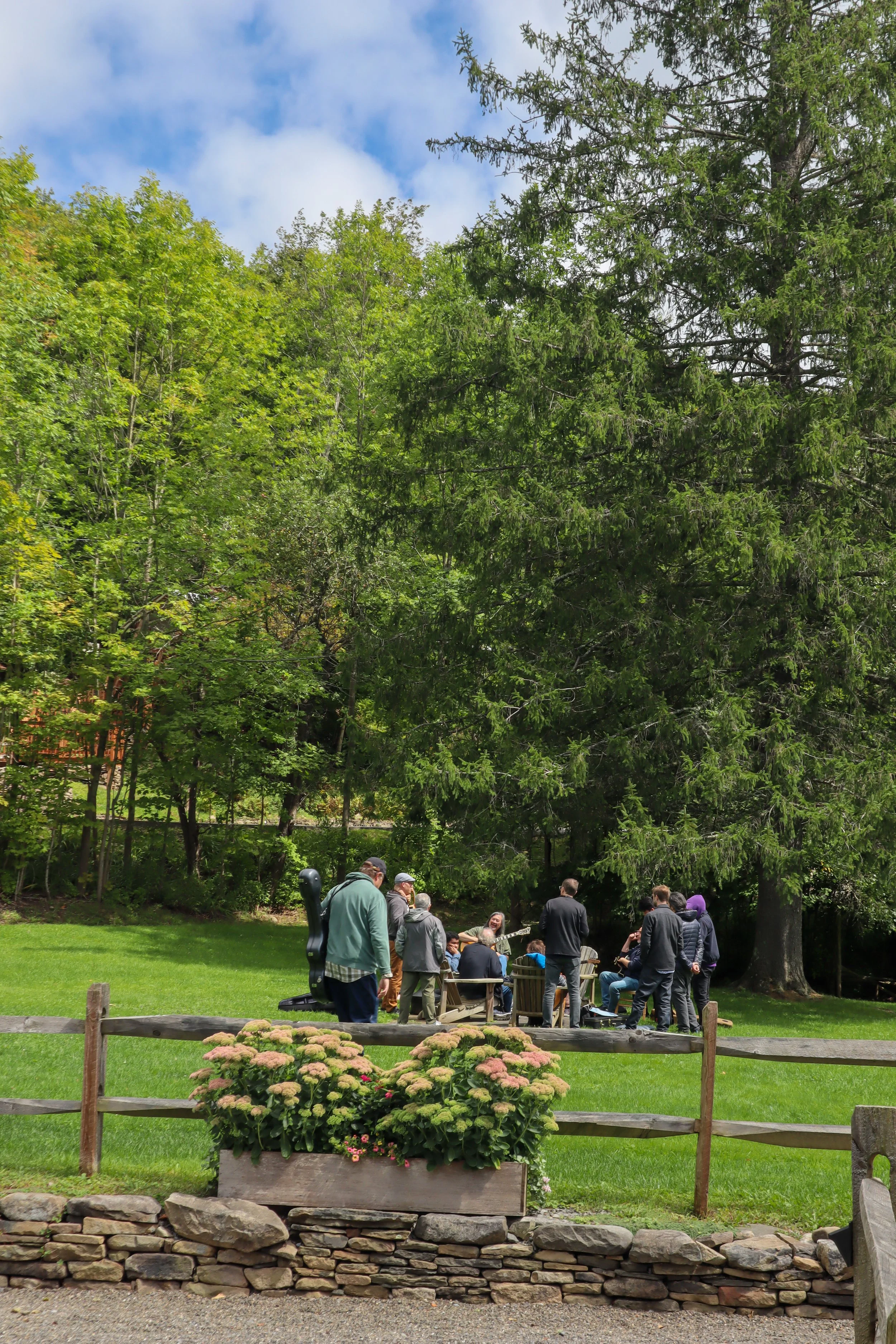 A group of people gathered outdoors in a park with green grass and large trees under a partly cloudy sky.