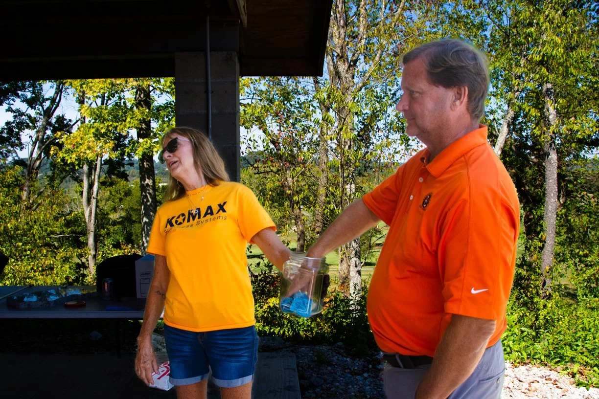 21st Annual Make-A-Wish Golf Tournament. A woman in a bright yellow t-shirt and sunglasses is laughing as a man in an orange polo shirt reaches into a jar. They are outdoors, surrounded by green trees and blue sky.