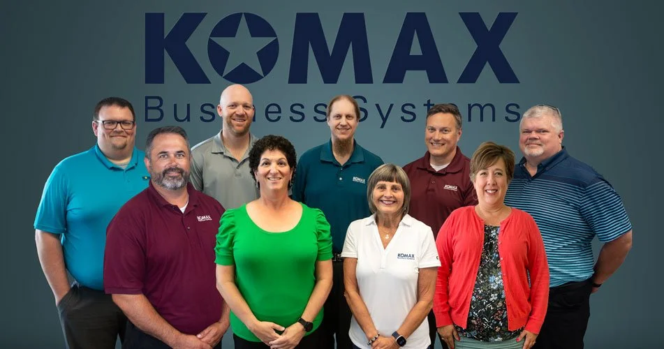 Group of nine people standing in front of a KOMAX Business Systems logo, posing for a photo inside an office.