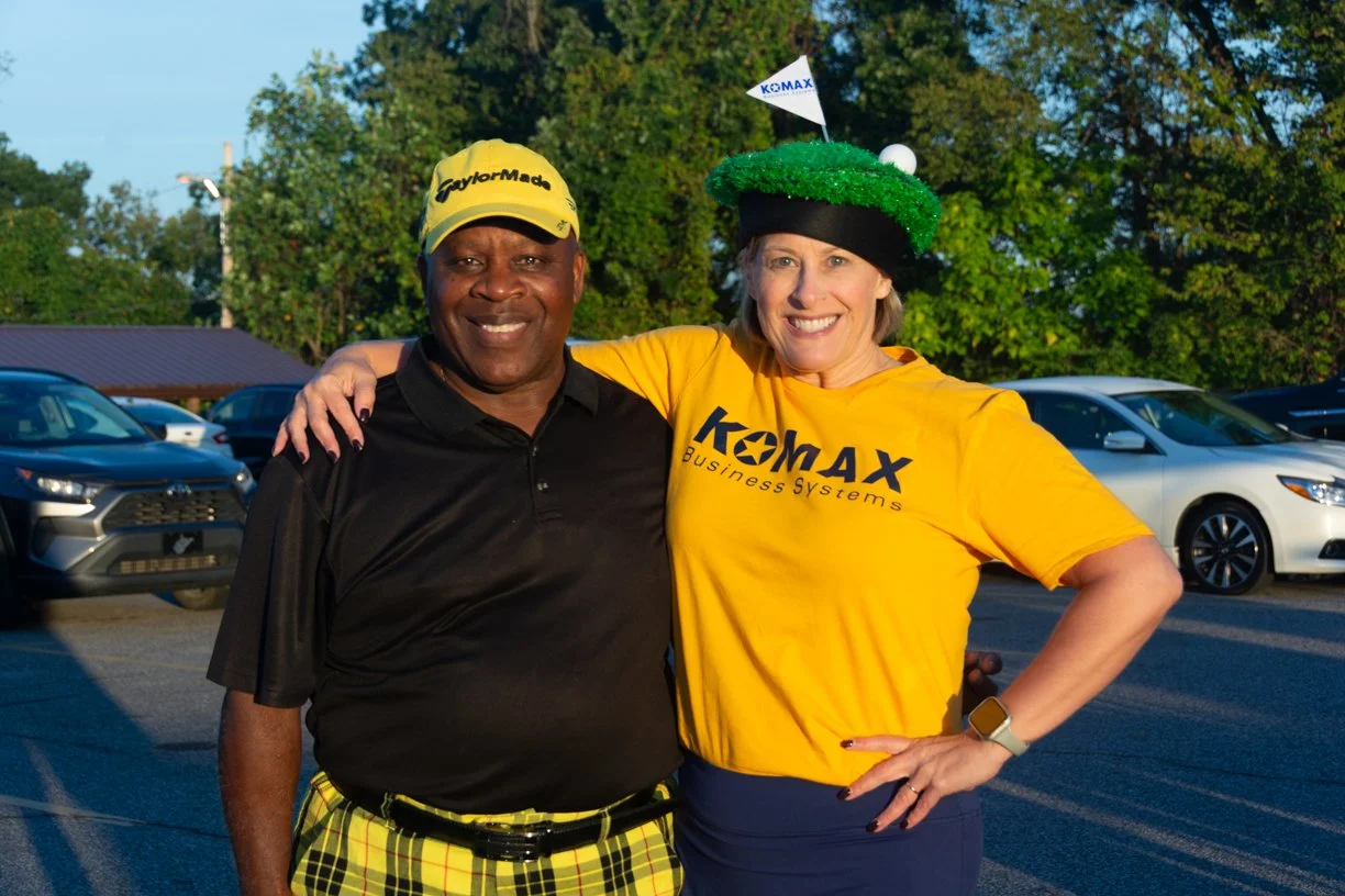 Two smiling people standing outdoors in parking lot, wearing yellow and black clothing with 'KOMAX' branding, one with a golf hat and the other with a festive green hat.