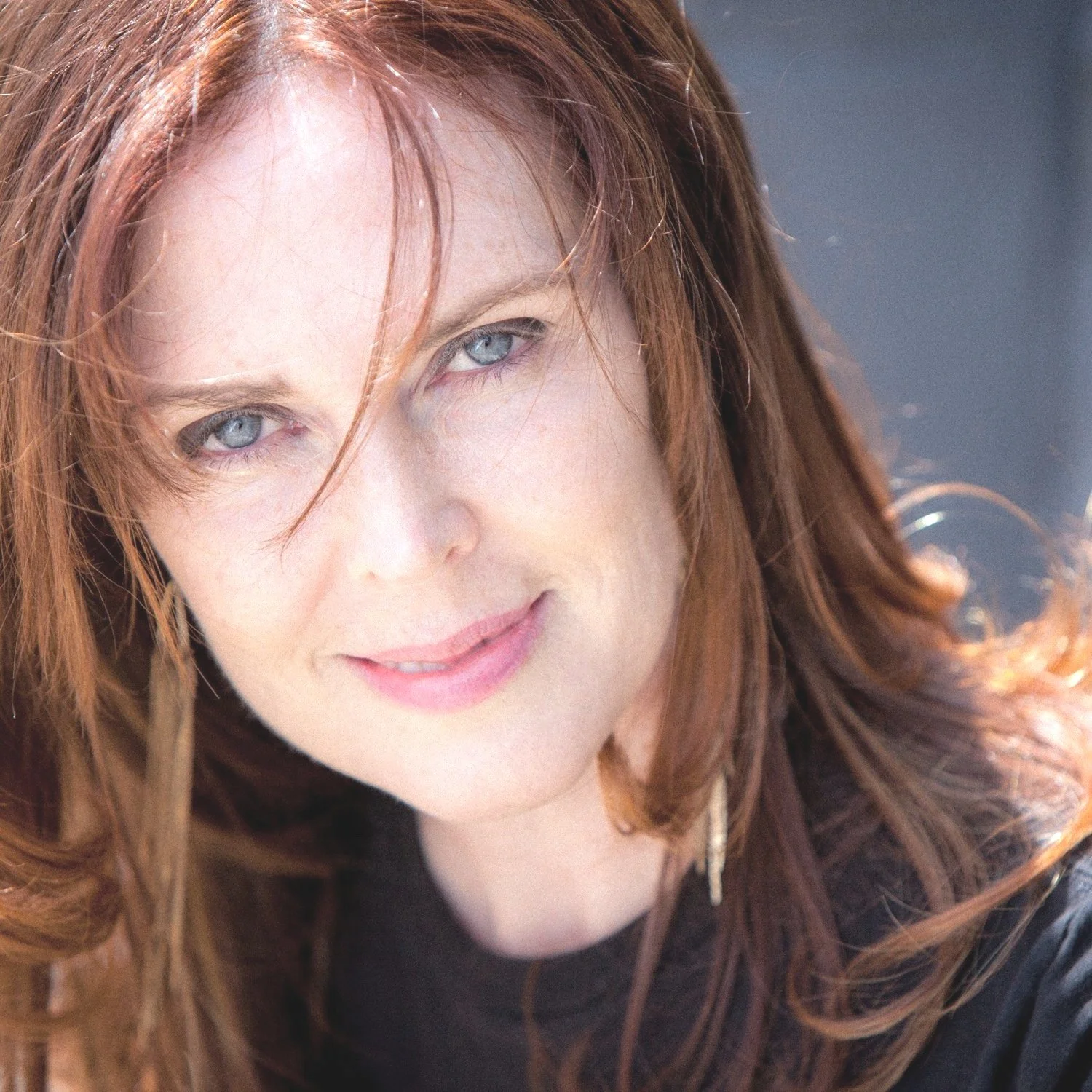A woman with long, wavy red hair and blue eyes looking at the camera, smiling slightly.