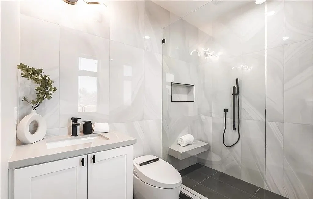 Modern bathroom with white cabinets, a shower area with glass doors, black shower fixtures, a countertop sink, green plant decor, and a toilet.