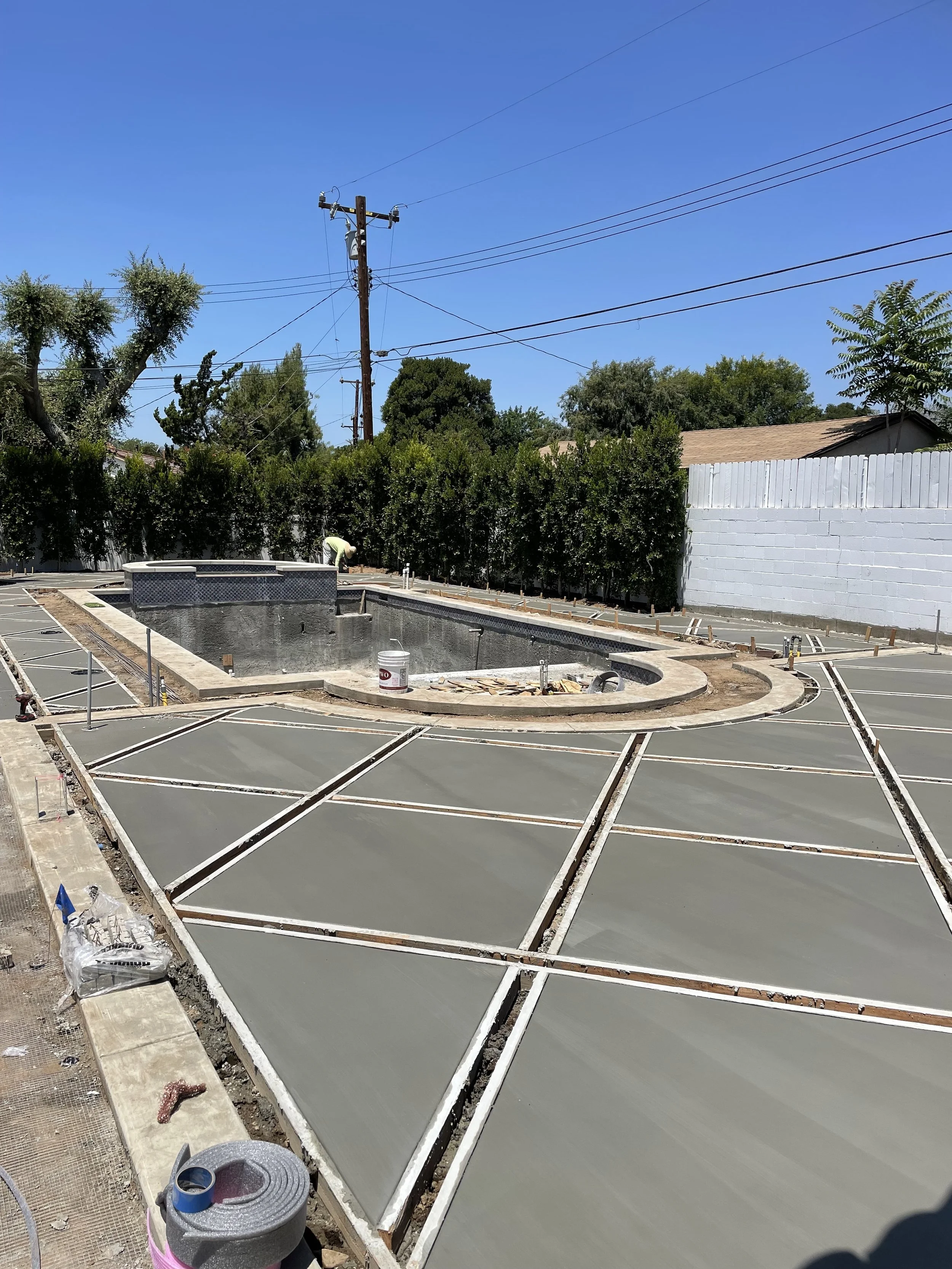 Construction site with a swimming pool under construction, concrete poured on surrounding slabs, and landscaping work ongoing, with trees and power lines in the background.