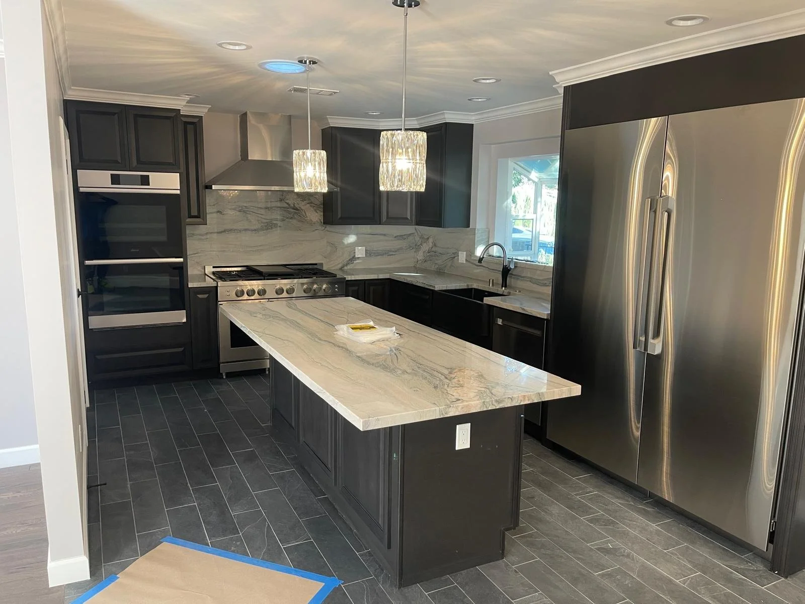 Modern kitchen with dark cabinetry, marble countertops and backsplash, large stainless steel refrigerator, oven, stove, and double pendant lights.
