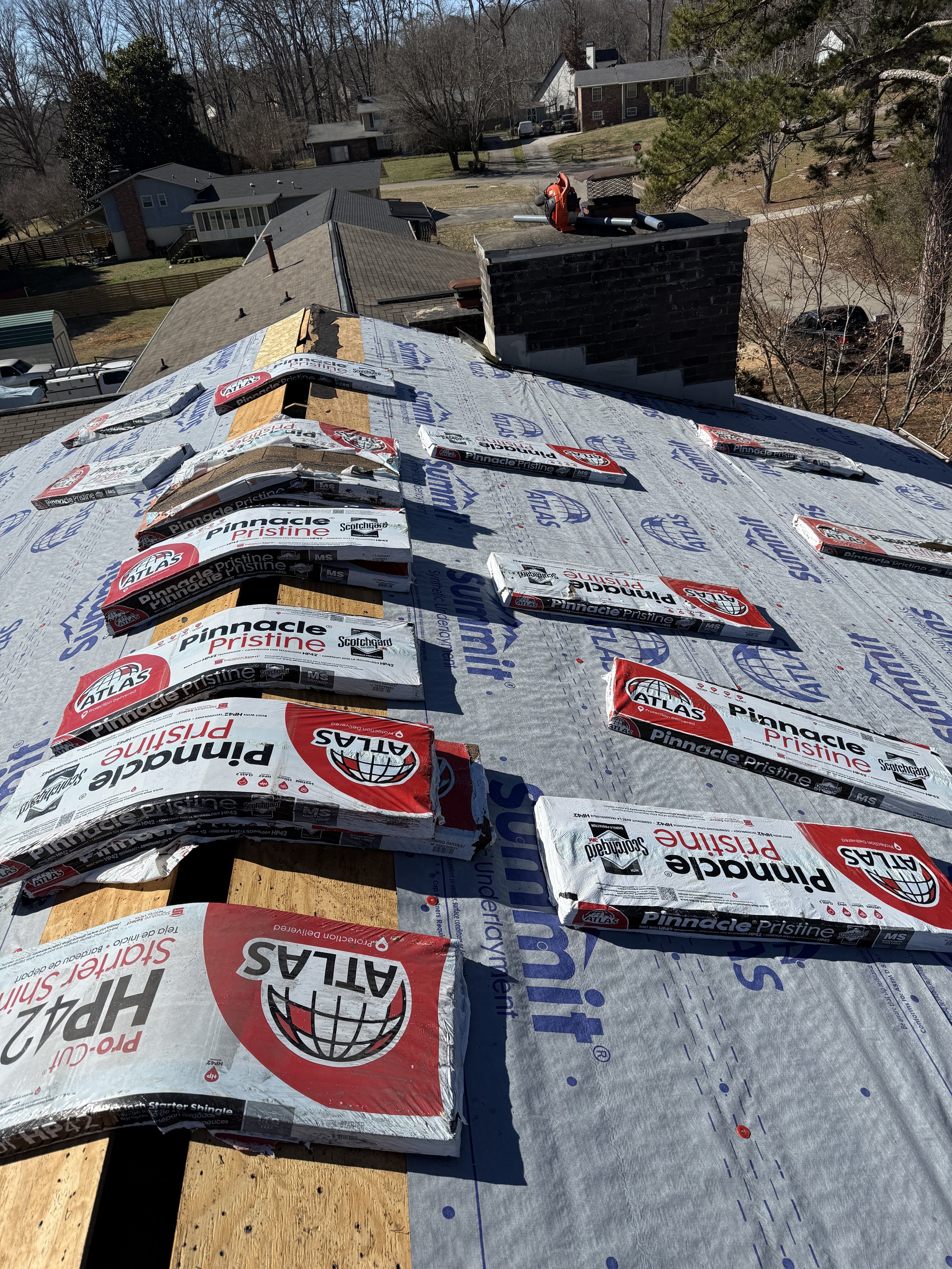 A rooftop under construction with bundles of atlas Pinnacle Pristine shingles placed on the roof, with neighboring houses and trees visible in the background on a sunny day.