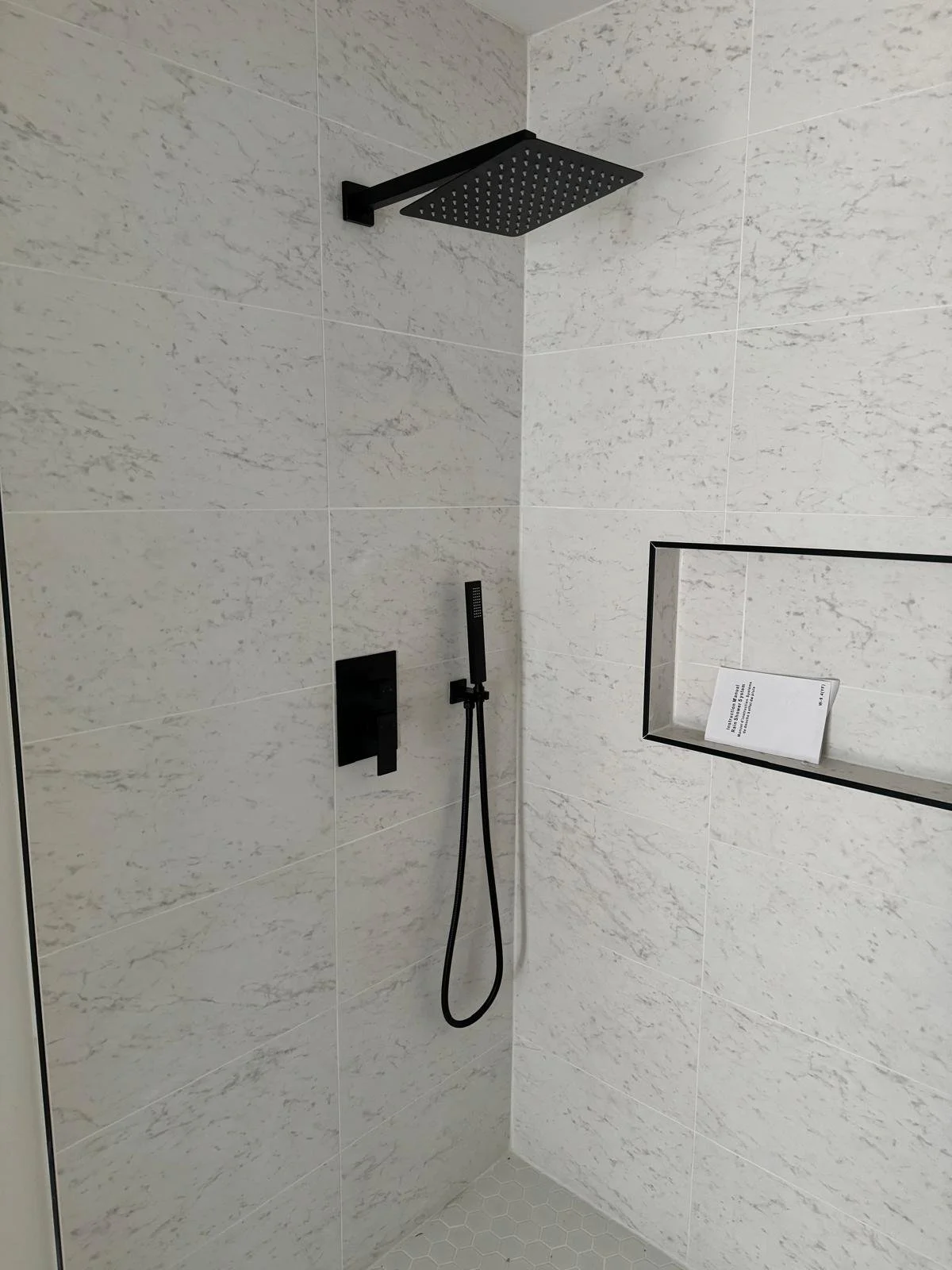 A modern shower with a black rainfall showerhead and a black handheld shower on a black wall mount. The shower walls are covered with white veined tiles, and there is a built-in niche with a book inside.