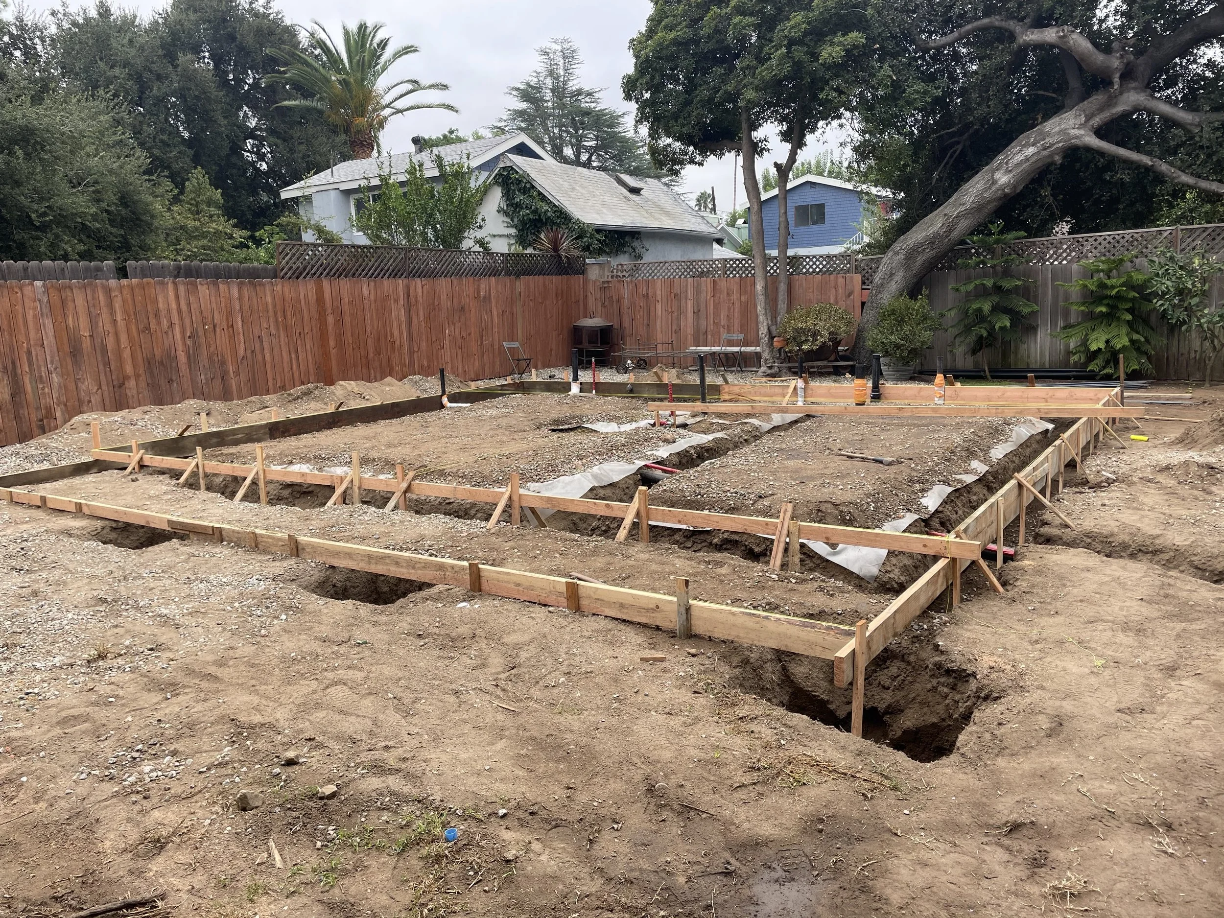 A backyard undergoing construction with wooden framing for a new structure, dirt, and construction tools, surrounded by a wooden fence and trees.