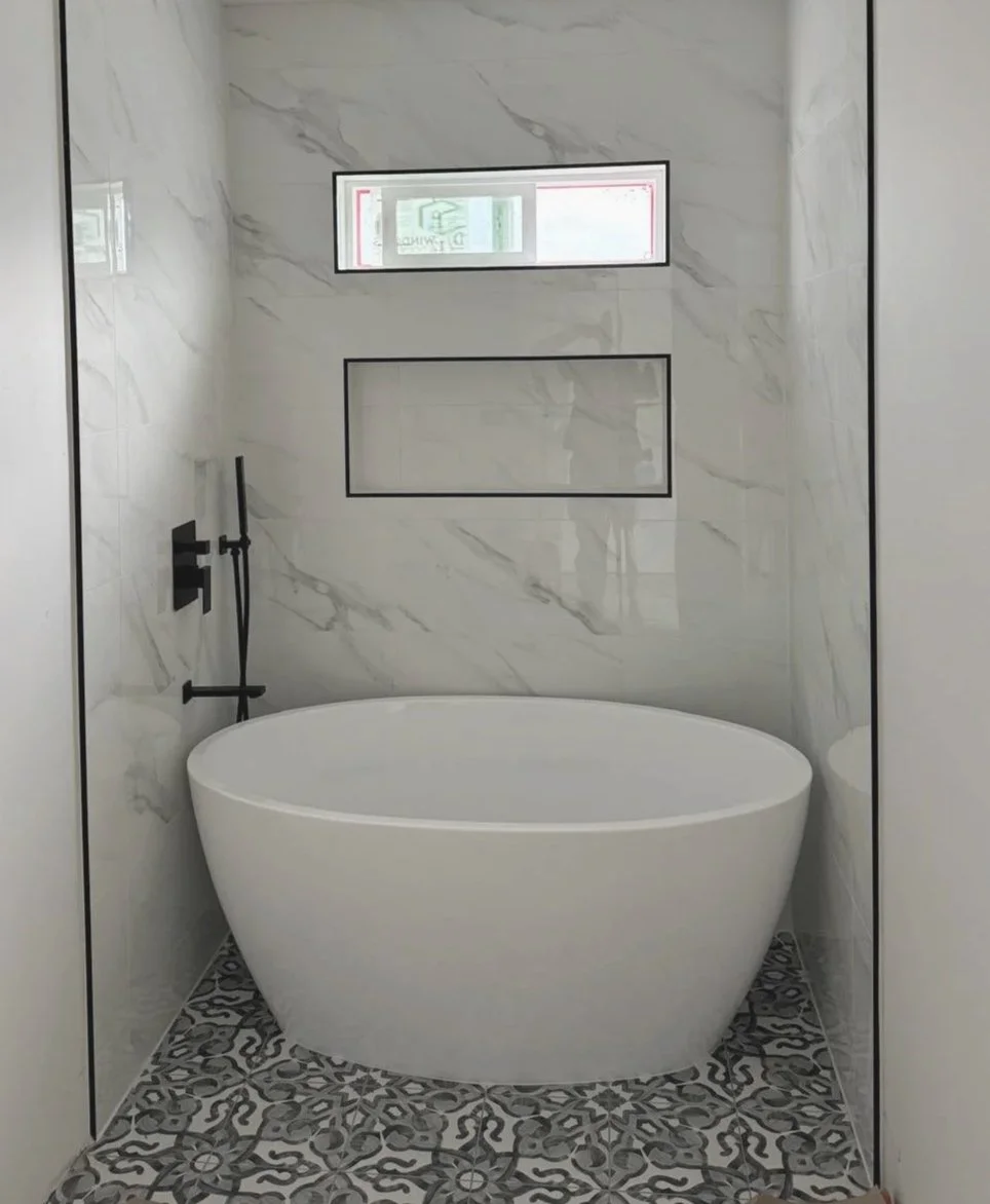 Modern bathroom with a freestanding oval bathtub, gray and white marble-tiled walls, a patterned tile floor, a narrow horizontal window, and black fixtures.