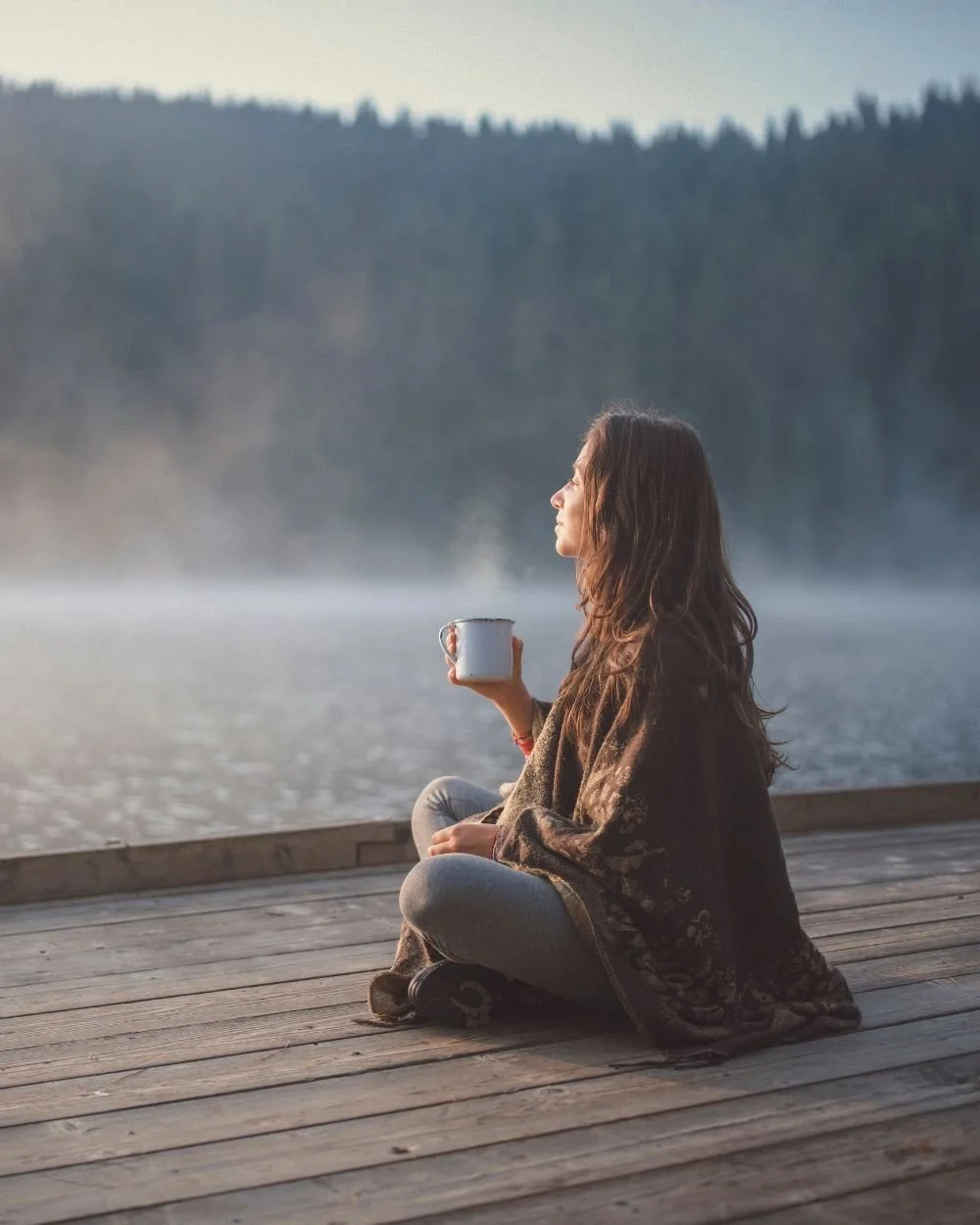 A woman sitting cross-legged on a wooden dock holding a mug, facing a foggy lake with forested mountains in the background.