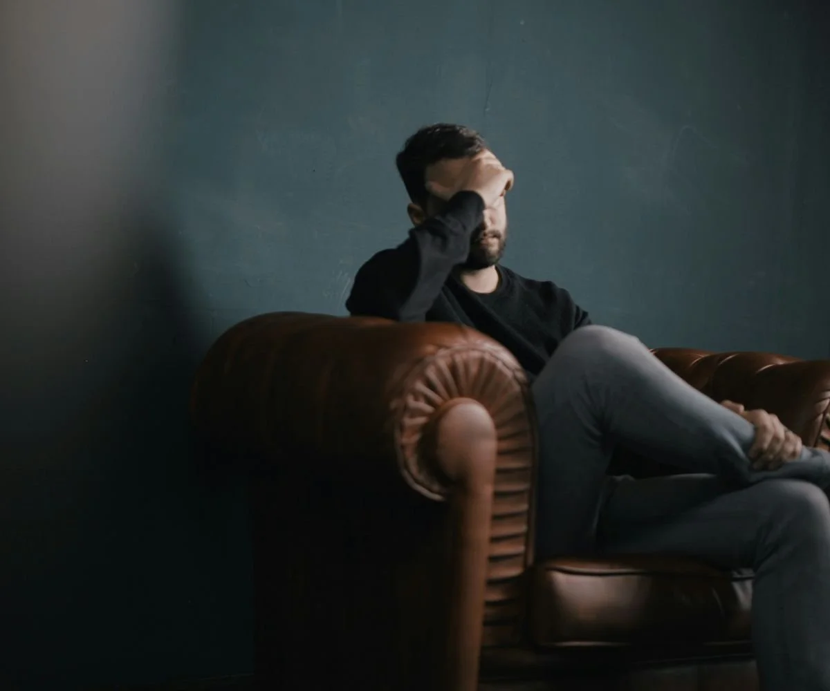 A man sitting on a brown leather couch, resting his forehead in his hand, in a dimly lit room with dark walls.