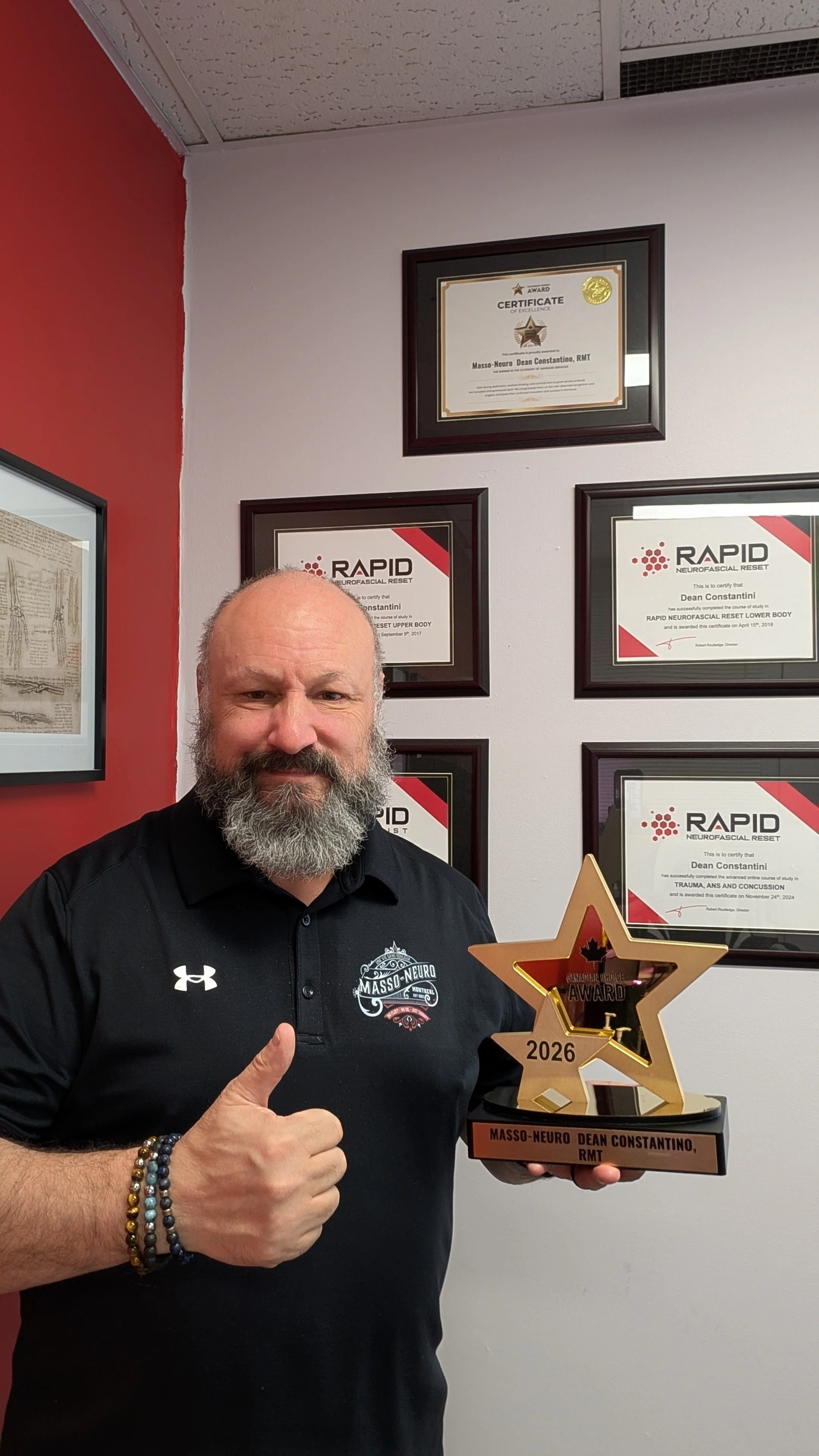 A man with a beard and a black shirt holding a star-shaped award and giving a thumbs-up. The wall behind him displays framed certificates and awards.