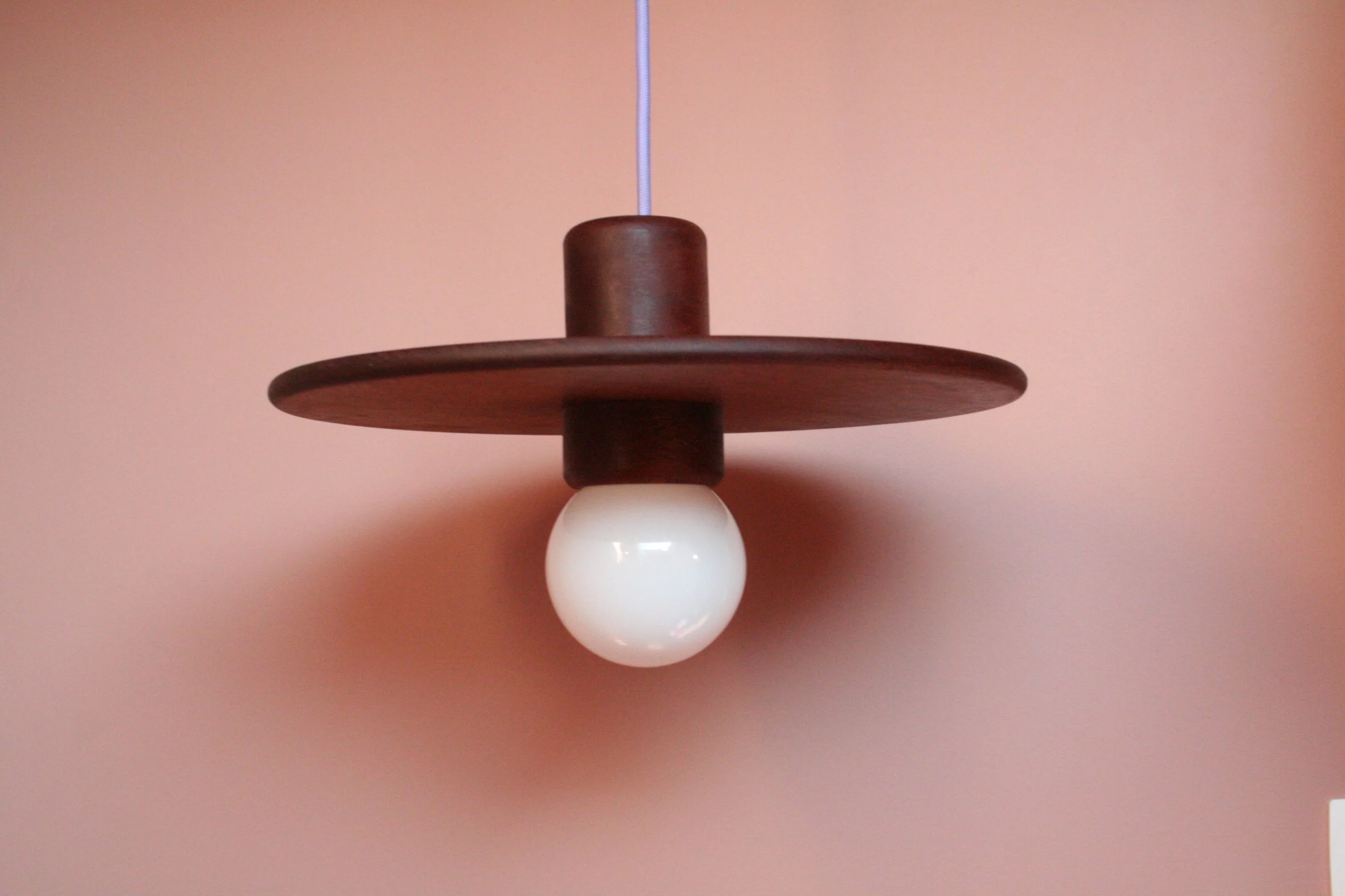 Houten plafondlamp met een witte ronde lampenkap, hangend aan een blauwe snoer, tegen een lichtroze muur.