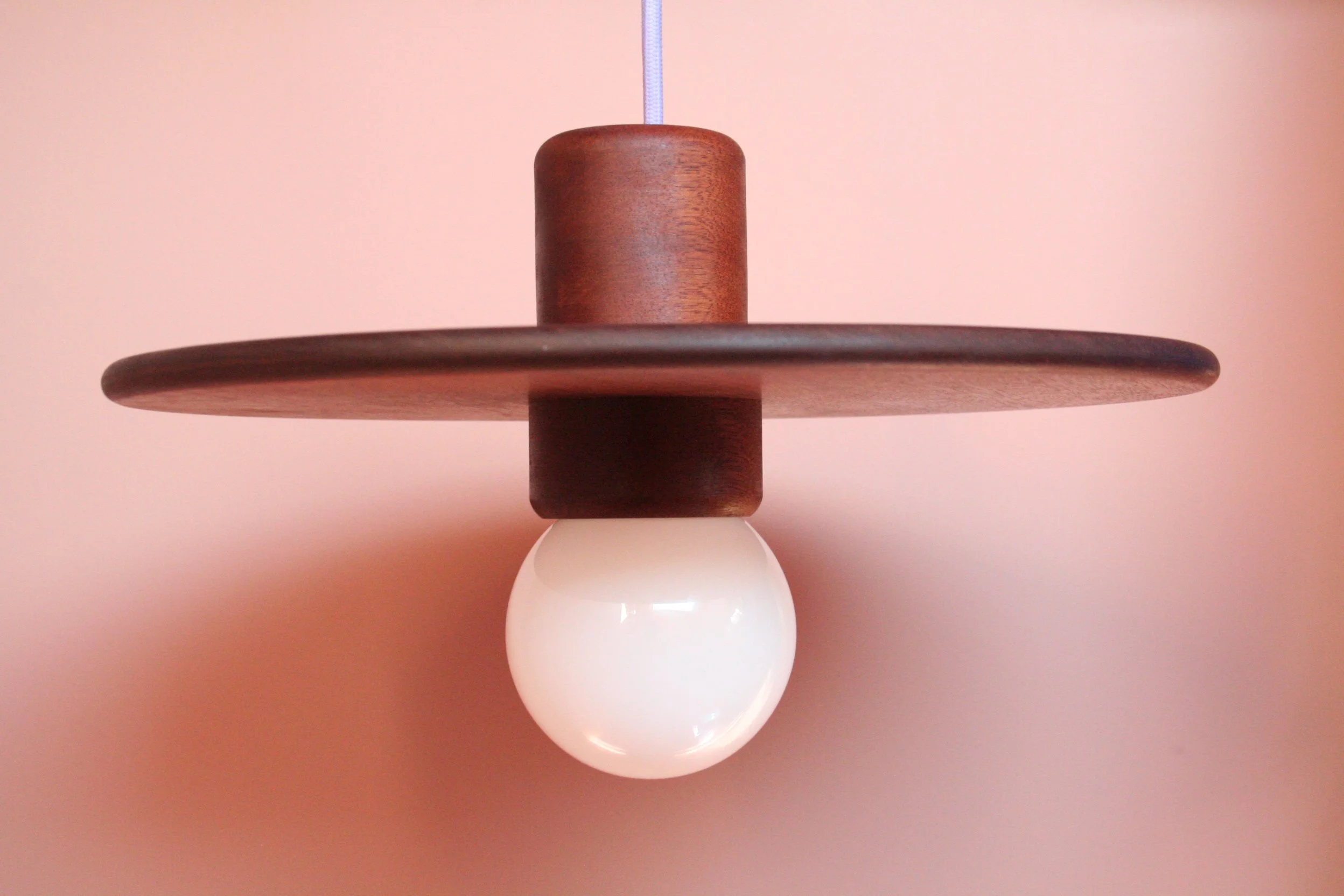 Een moderne plafondlamp met een witte glazen bol, houten en metalen elementen, hangend aan een blauwe draad, tegen een lichtroze muur.