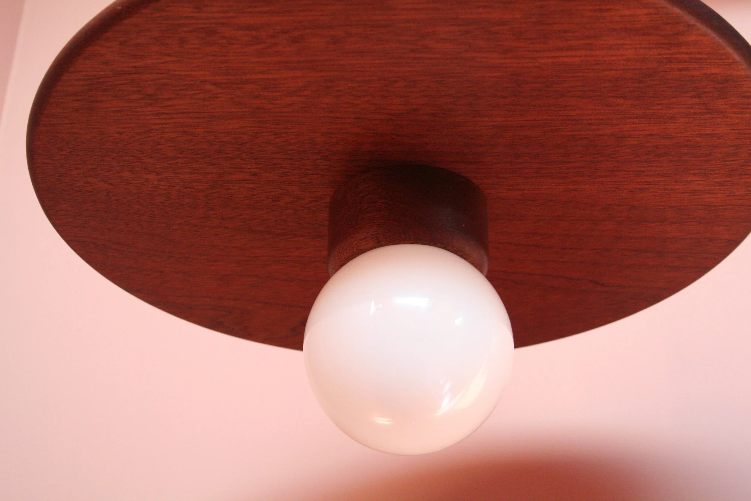 Close-up van een plafondlamp met een ronde witte bol op een houten plafond tegen een lichtroze muur.