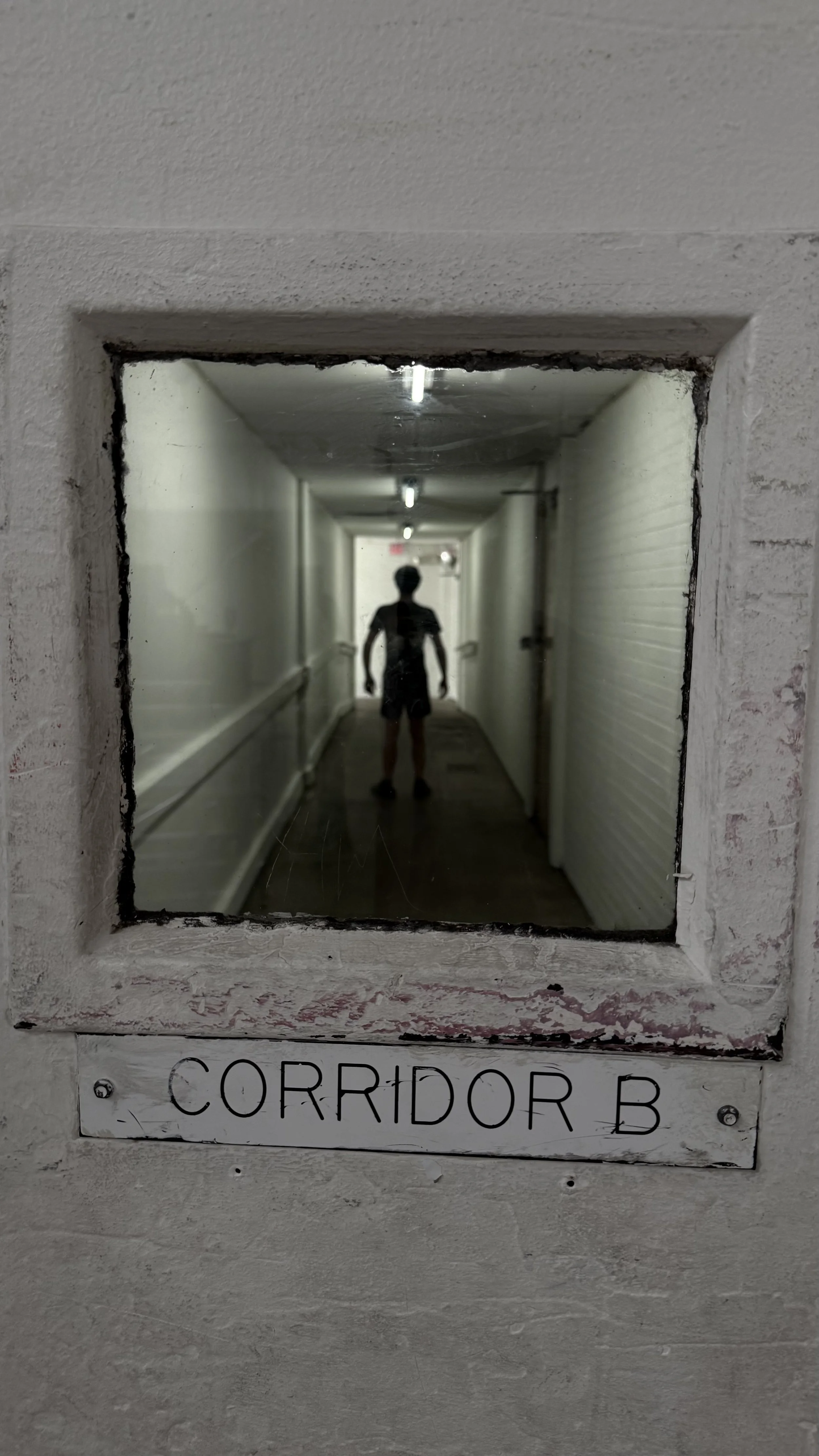 A person standing at the end of a narrow, dimly lit corridor labeled 'Corridor B.' The photo is taken through a window looking into the hallway.