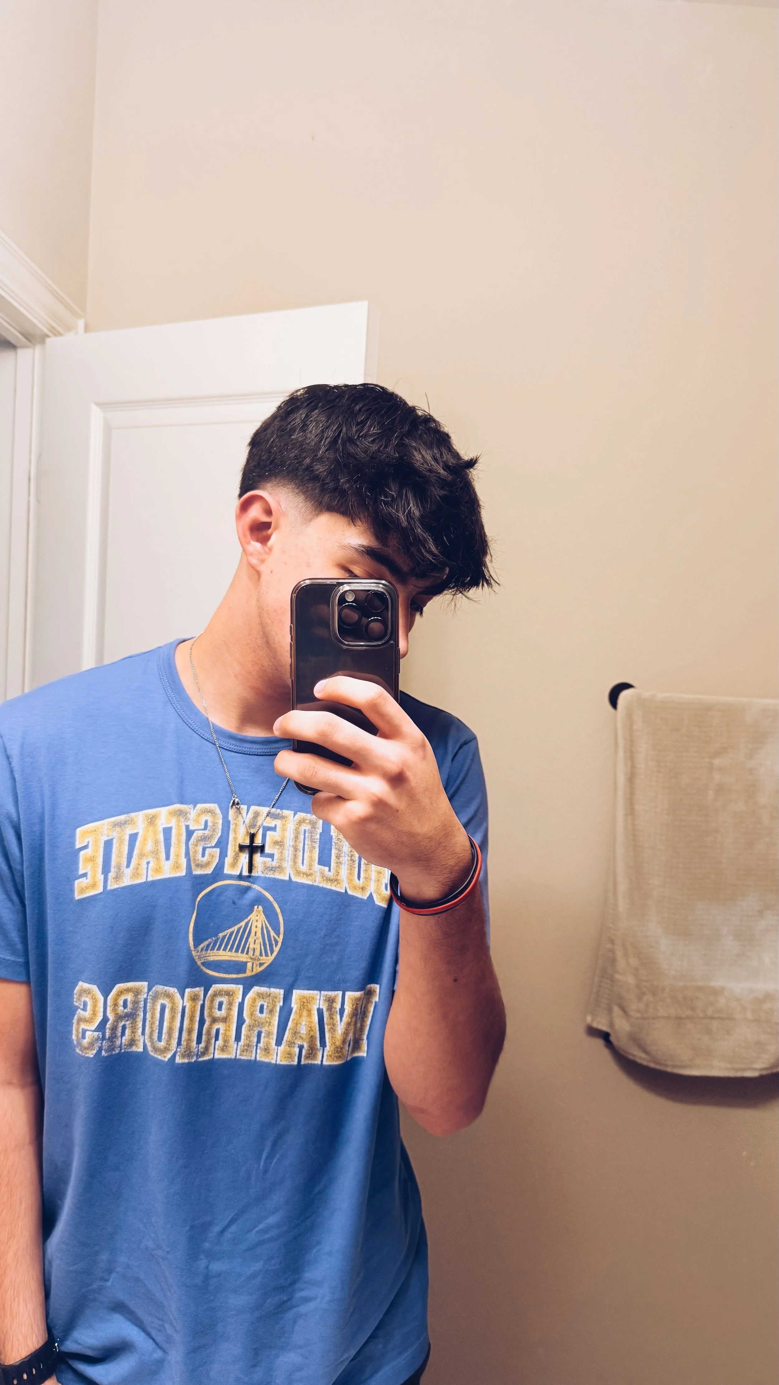 Person taking a selfie in a mirror wearing a blue Golden State Warriors shirt, holding a smartphone to their face, with a towel hanging in the background.