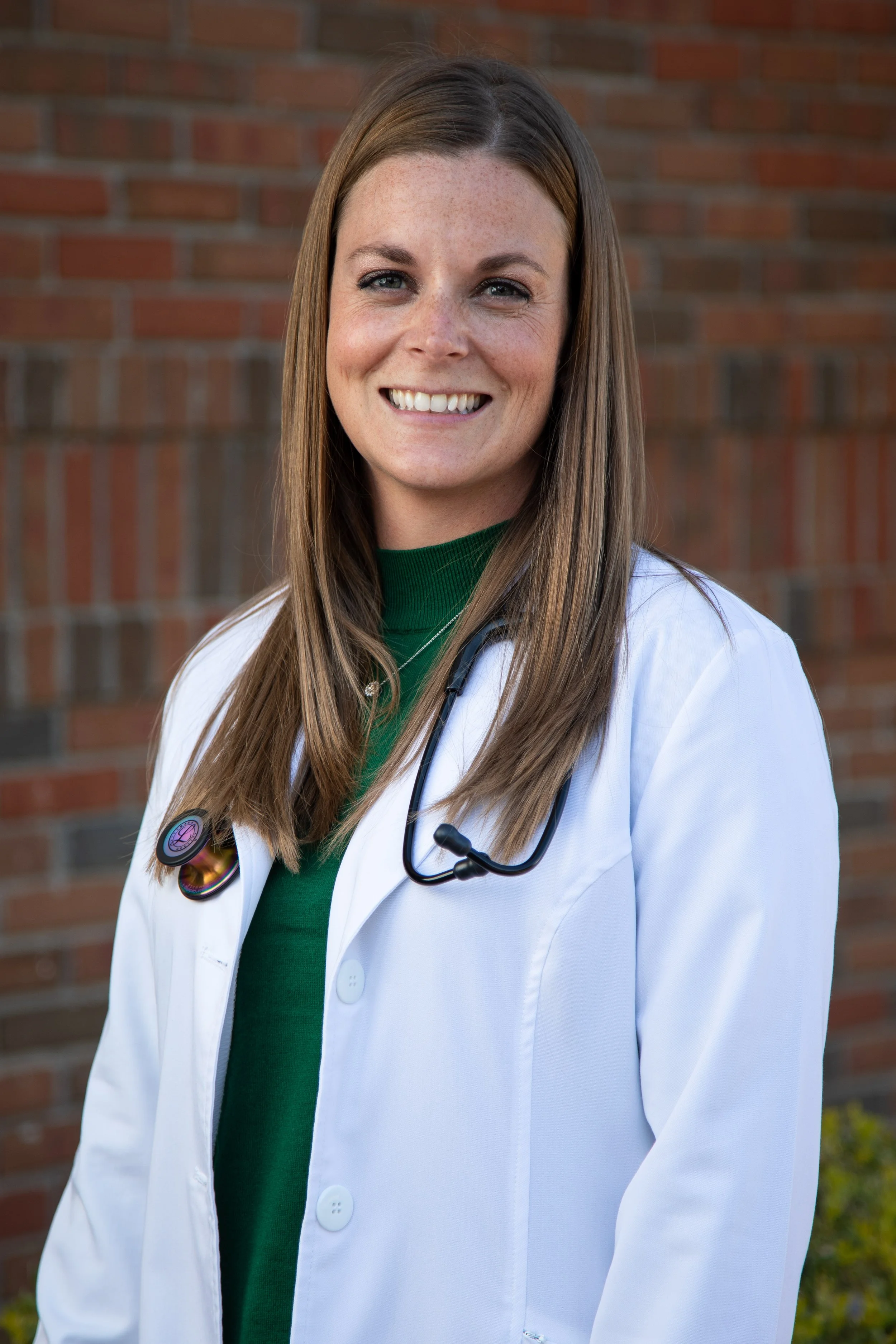 A smiling female healthcare professional with long brown hair wearing a white lab coat and green shirt, with a stethoscope around her neck, standing outdoors in front of a brick wall.