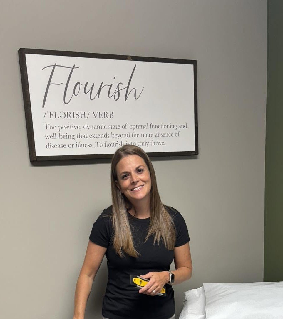A woman smiling and standing next to a horizontal white sign that reads 'Flourish' and defines it as a verb meaning a positive, dynamic state of optimal functioning and well-being that extends beyond the absence of disease or illness.