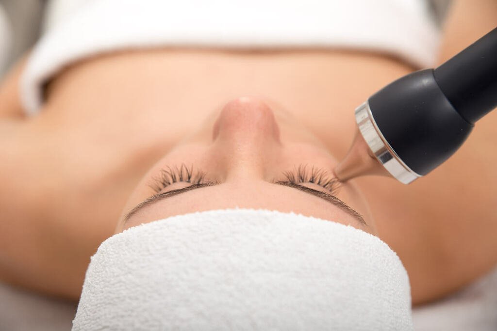 Personne recevant un traitement facial avec un dispositif cosmétique pour la peau du visage, tête enveloppée dans une serviette.