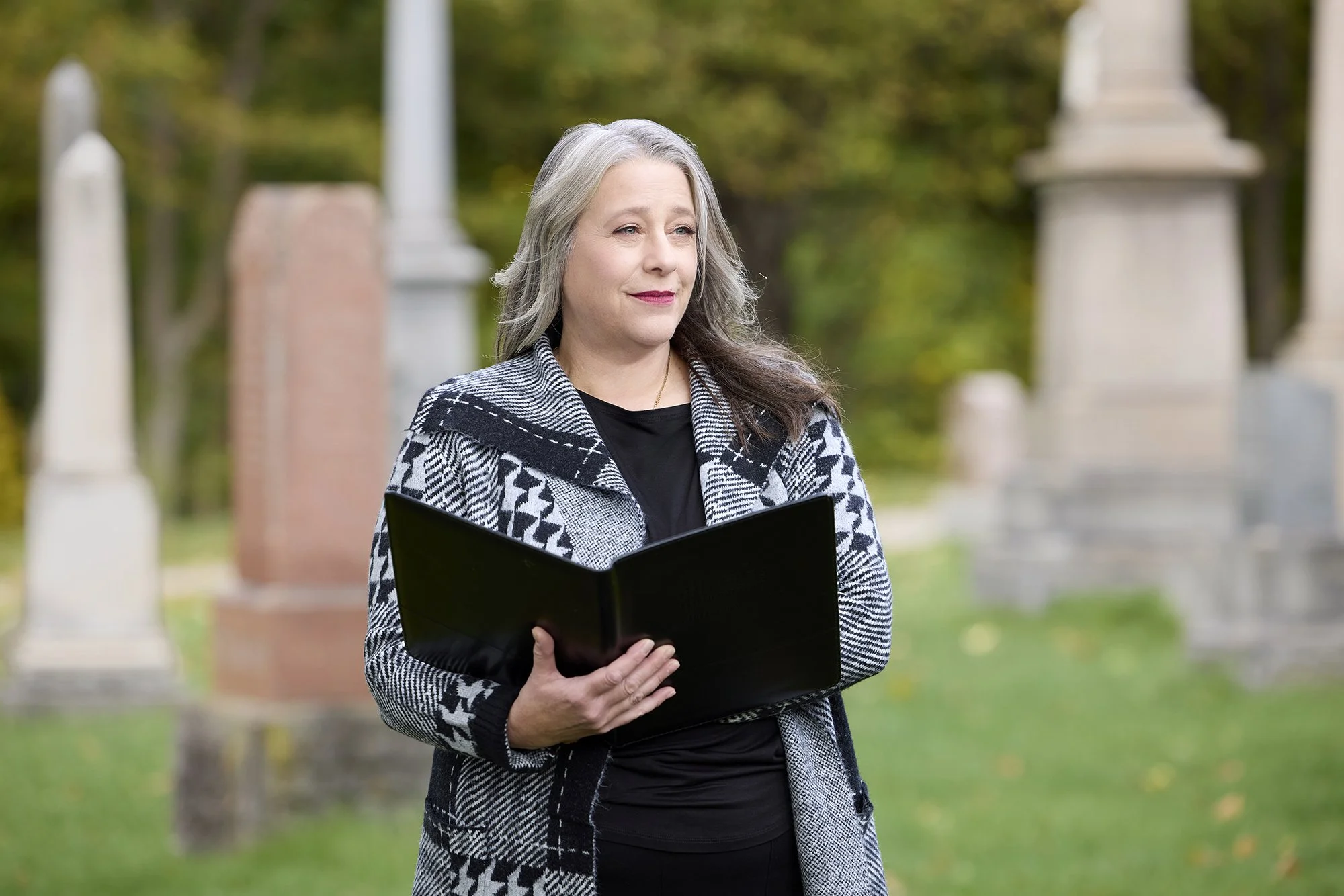 Jeanne-Louise holding an open black book, standing outdoors near several tombstones in a cemetery with green trees in the background.