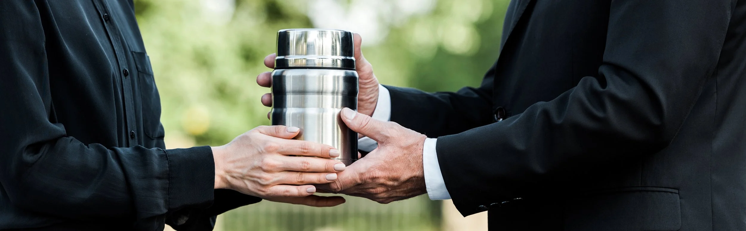 Two people in formal attire exchanging a stainless steel thermos outdoors.
