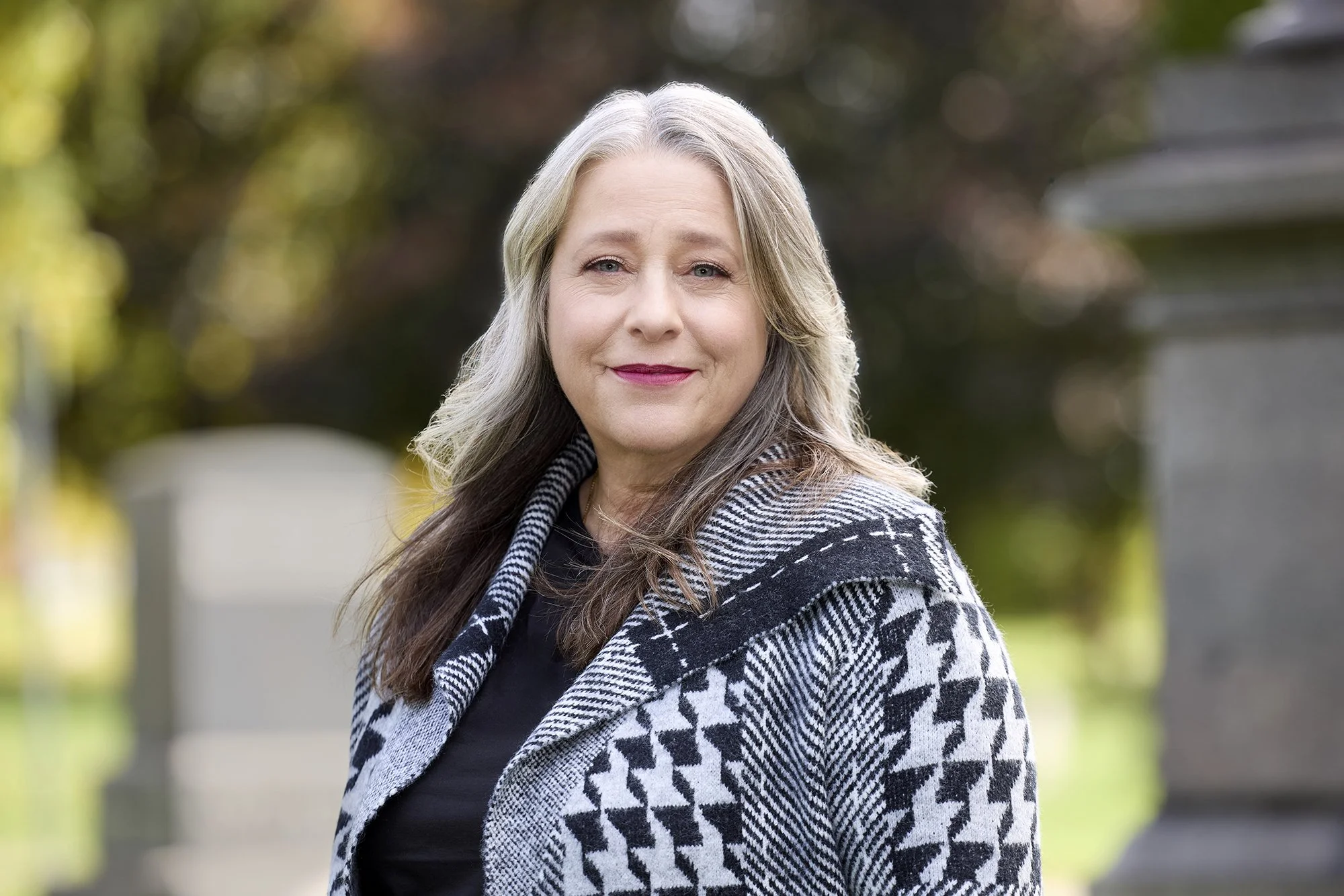 A woman with gray hair, wearing a black top and a black and white houndstooth patterned coat, standing outdoors with trees and a stone structure in the background.
