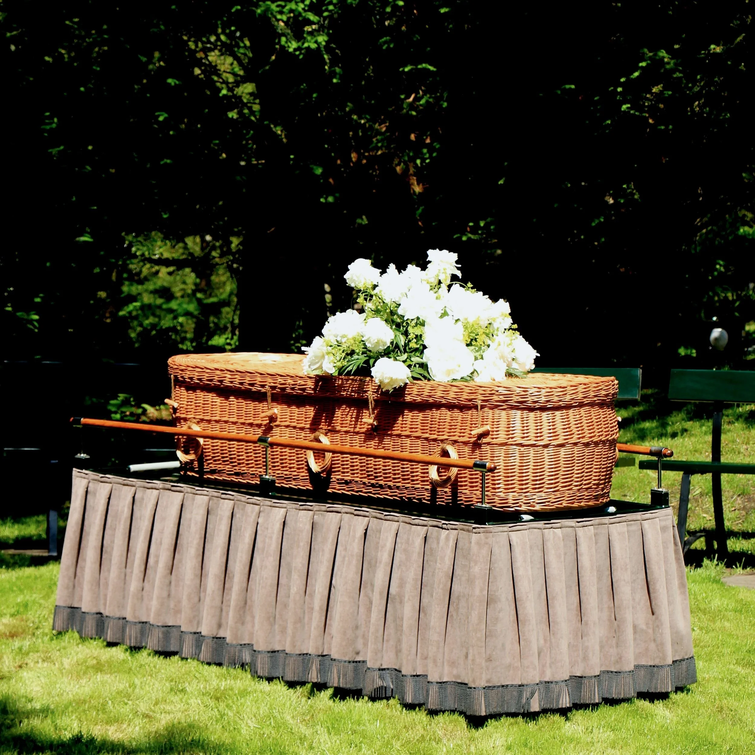 A casket placed on a decorated black table outdoors, with white flowers on top and a beige skirted covering, set in a sunny garden area with trees in the background.