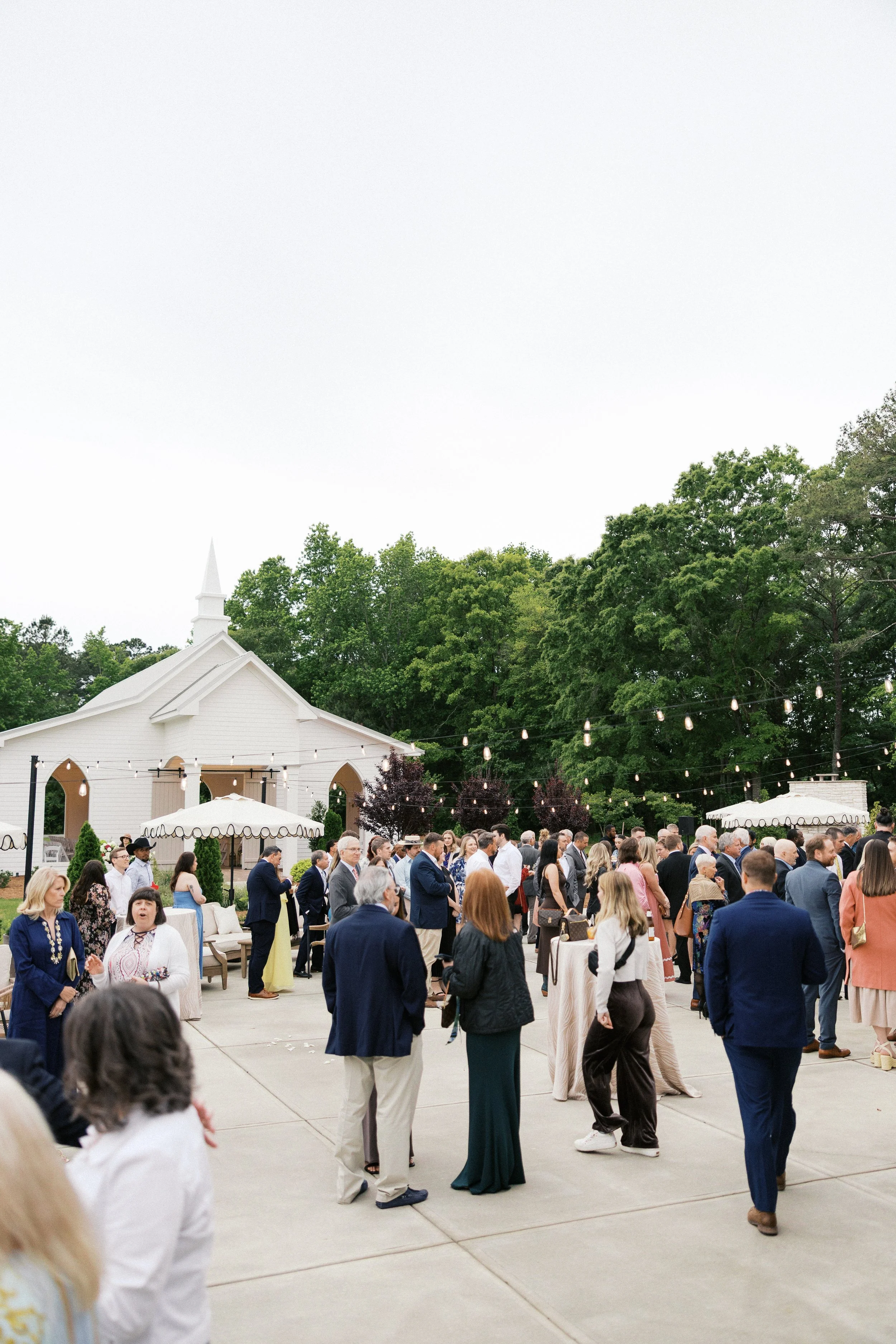 Outdoor patio for cocktail hour with seating, umbrellas, and string lights in front of a white house with trees in the background.