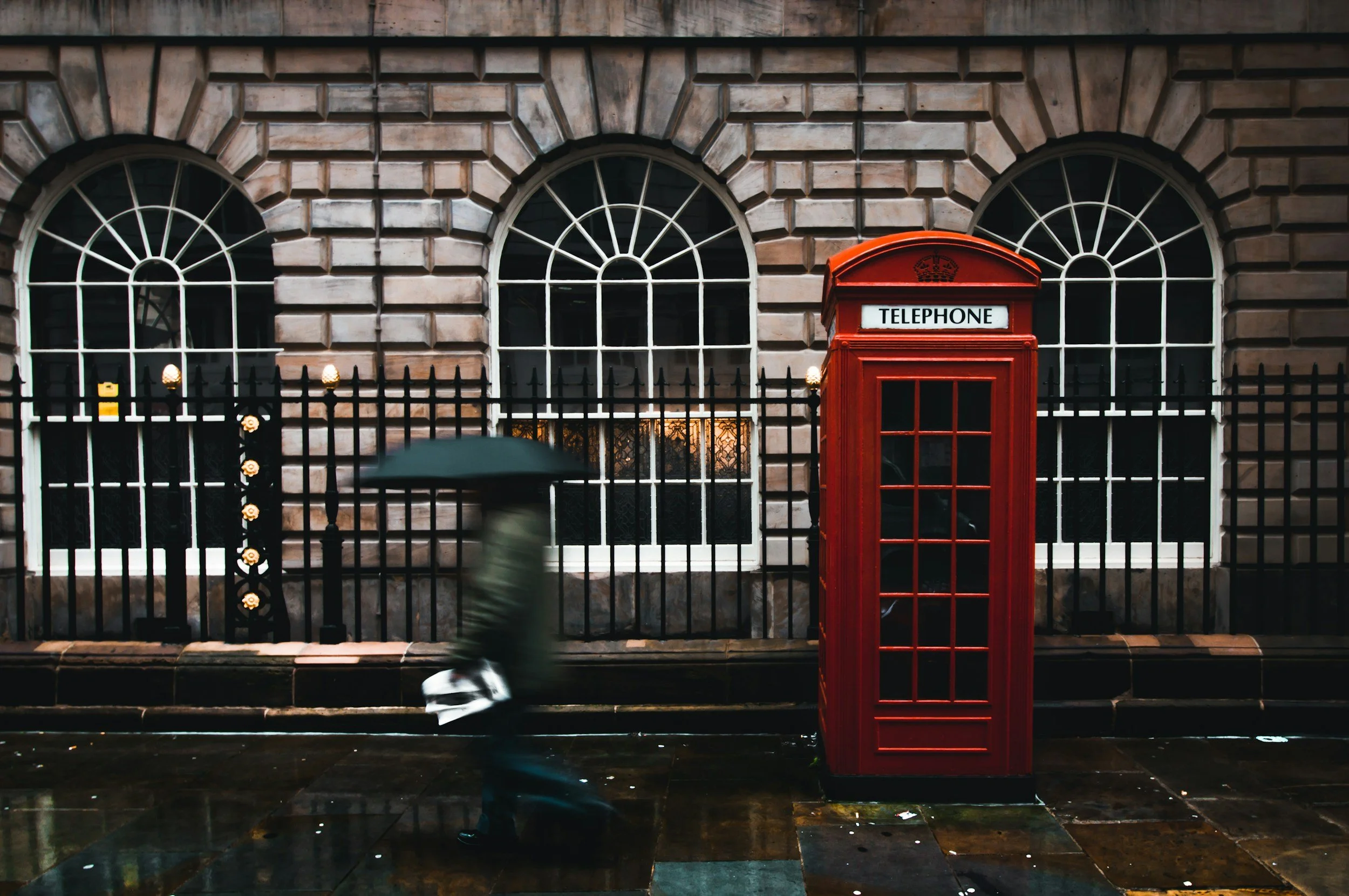 Red British telephone booth, rainy street, person with umbrella, arched windows, stone building, wrought iron fence.