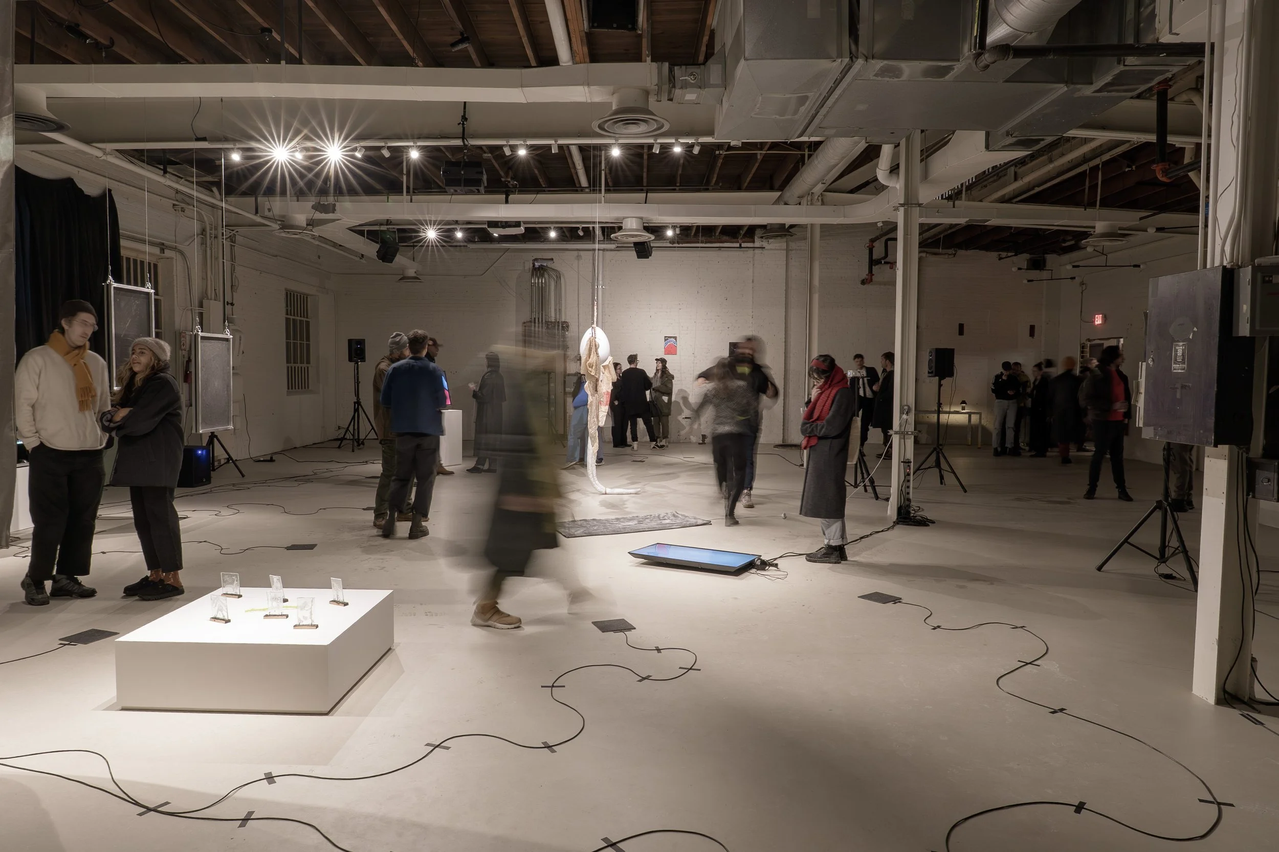 An open warehouse style room with light grey floor and walls with spotlit art and digital technology art installation with people moving between the pieces.