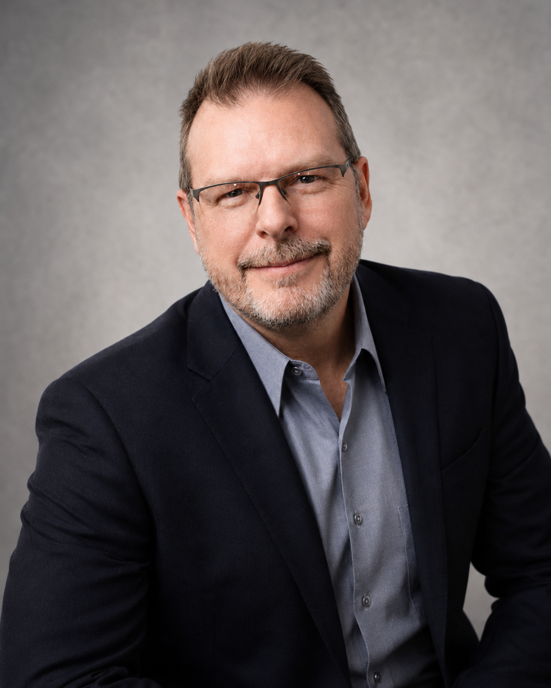 Man wearing glasses and a suit looks at the camera against a plain background.