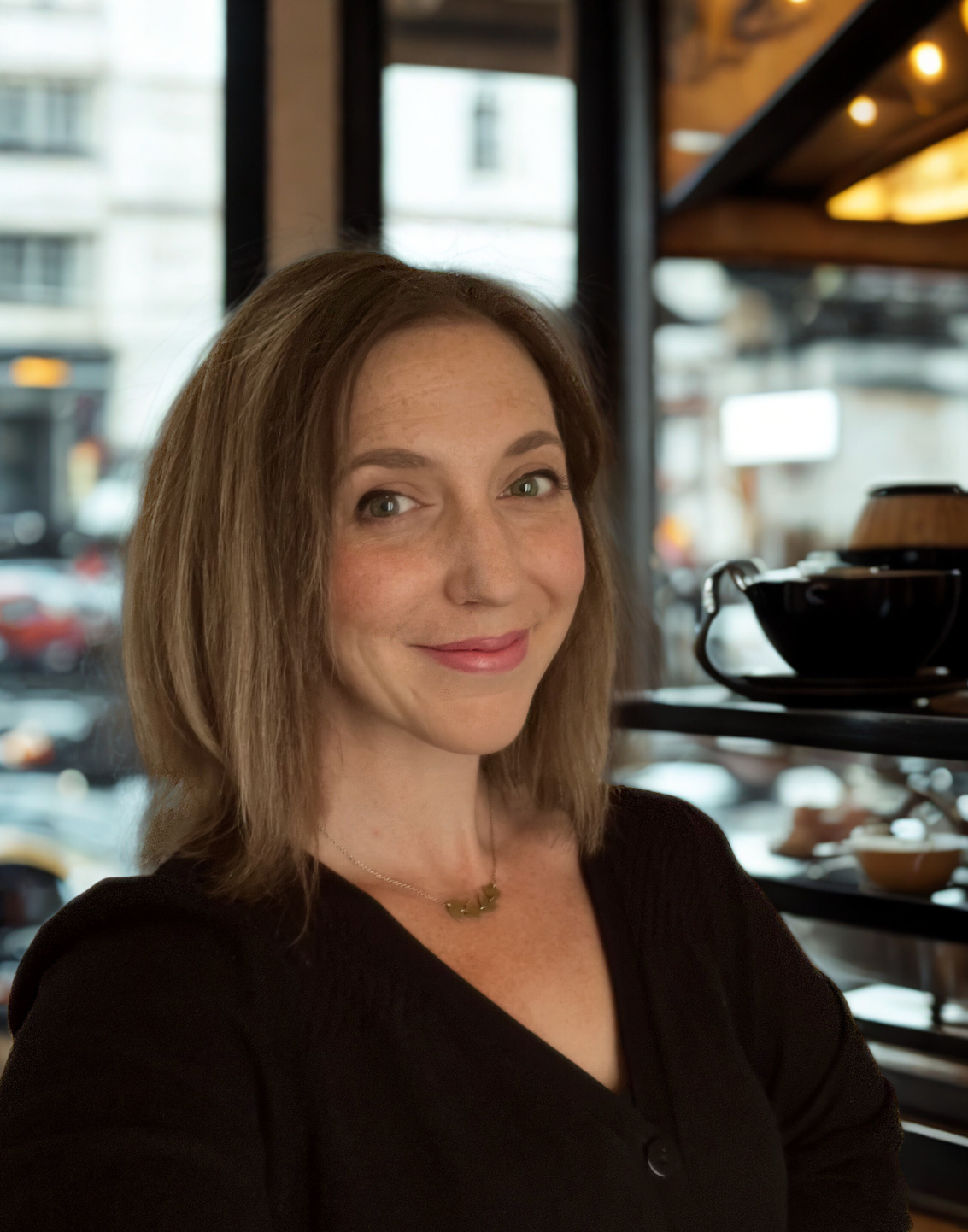 Mary Griffin Virtual Assistant, a woman with shoulder-length light brown hair smiles while sitting in a cafe, with a window showing parked cars and buildings outside.