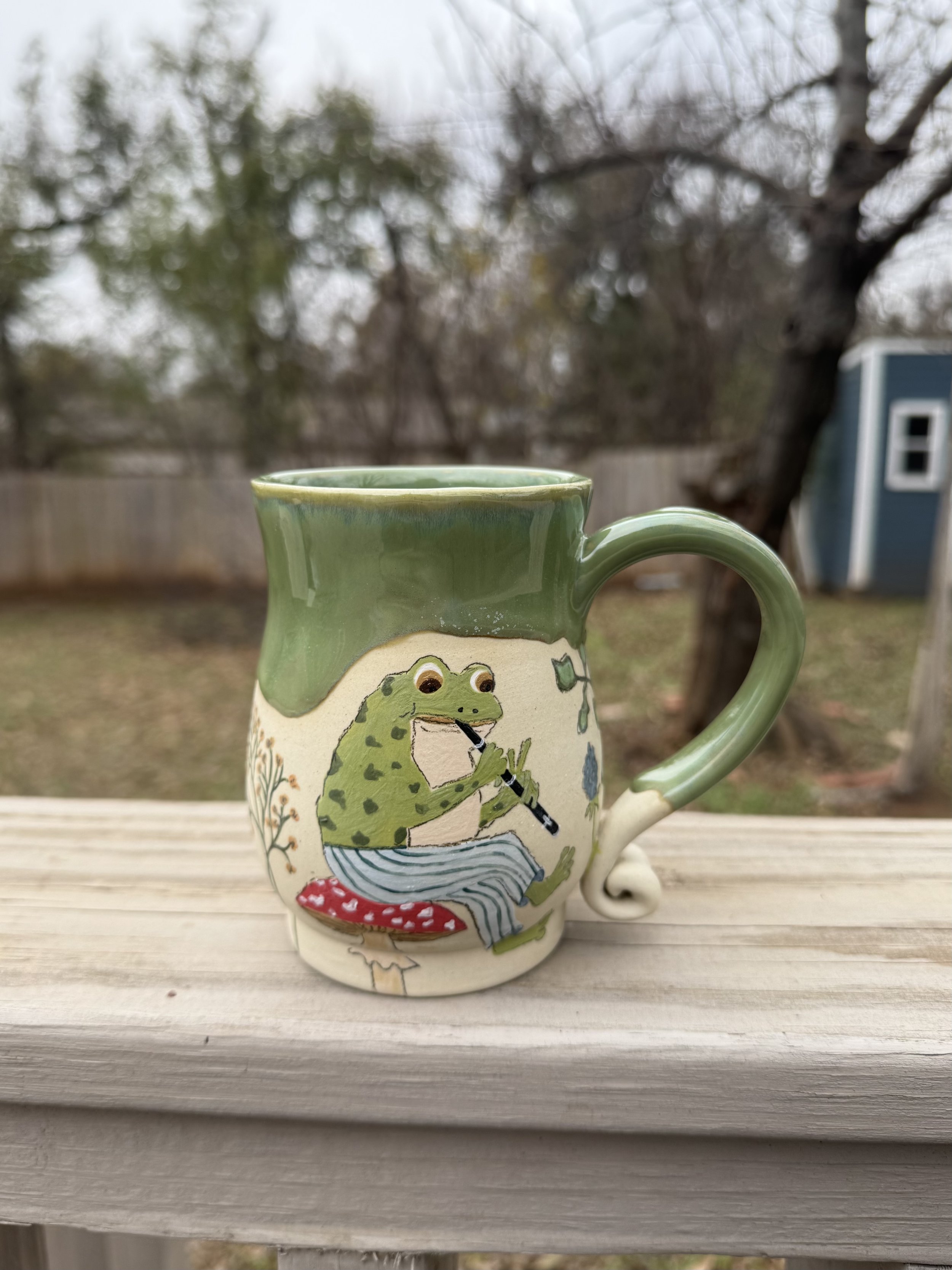 Ceramic mug with green glaze on top and a frog sitting on a toadstool playing a flute on the bottom