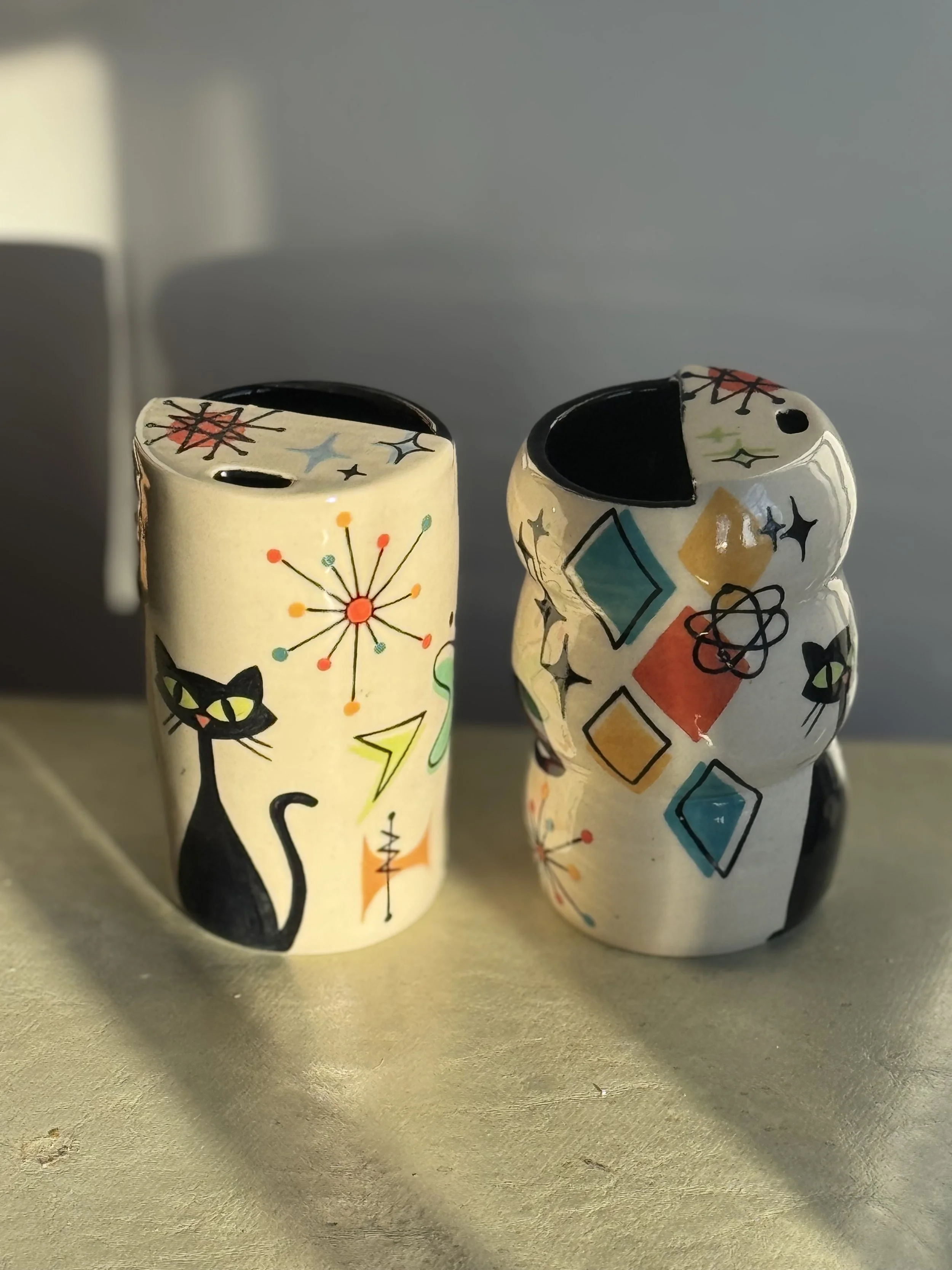 Two ceramic travel mugs with geometric shapes and a cat in bright colors on white with black outlines