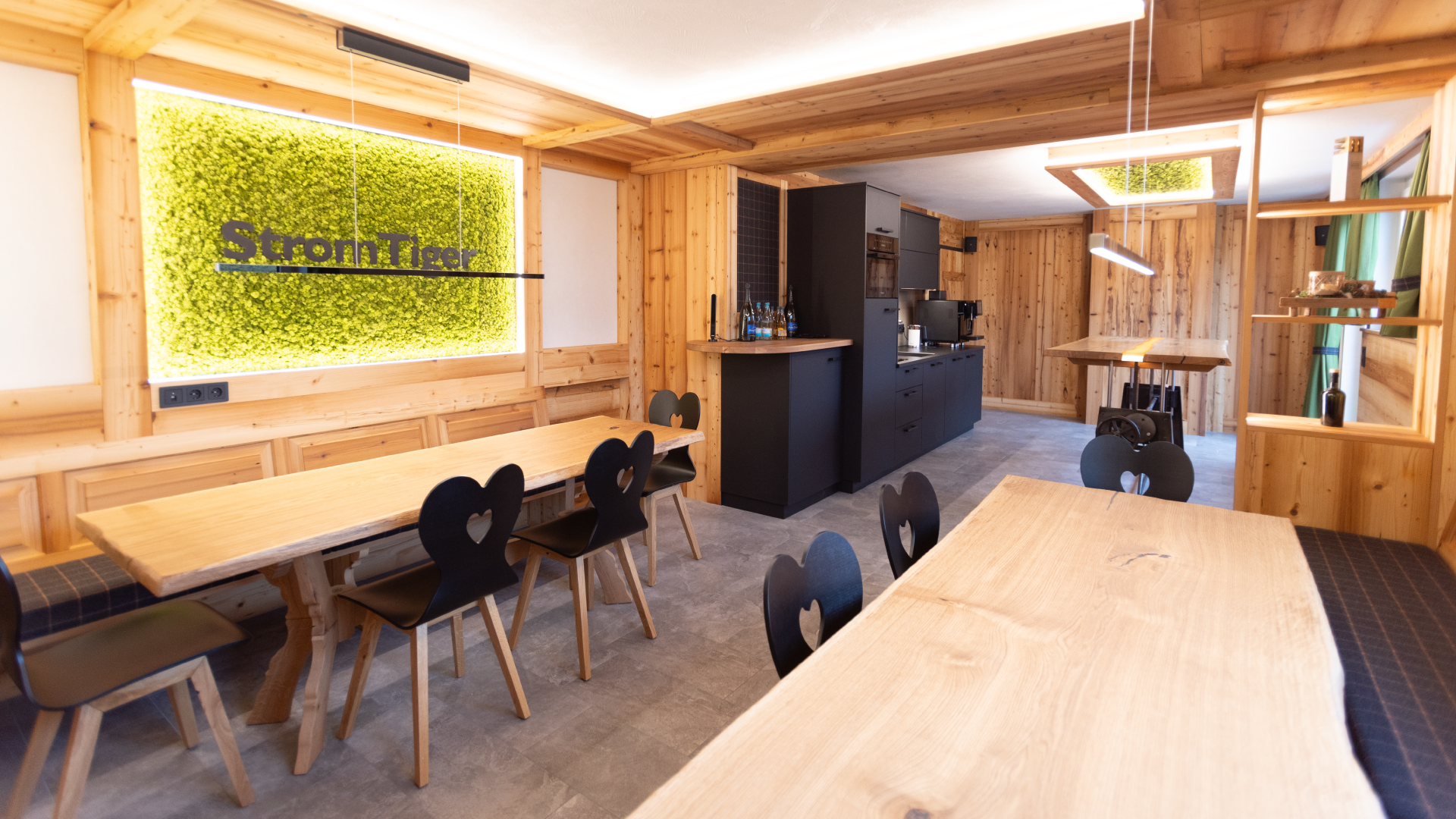 Innenansicht eines gemütlichen Raumes mit Holzvertäfelung, Tischen und schwarzen Stühlen, einem schwarzen Küchenblock, und einer Wand mit begrüntem Kunstwerk mit der Aufschrift 'StrongTiger'.