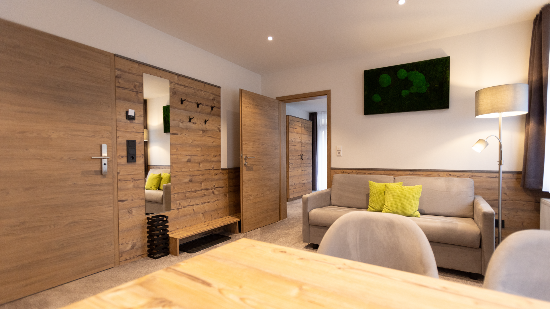 Gemütliches Wohnzimmer mit grauem Sofa, grünem Kissen, Holzpaneelen an den Wänden, Stehlampe, Wandbild mit grünen Punkten, Holzstuhl, Holztisch, offener Tür zum Schlafzimmer mit weiteren grünen Kissen auf dem Bett.
