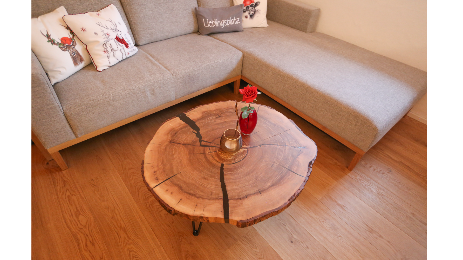Wohnzimmer mit einem grauen Sofa, dekoriert mit Weihnachtskissen, und einem runden Holztisch mit einer roten Rose in einer Vase und einem Kerzenhalter.