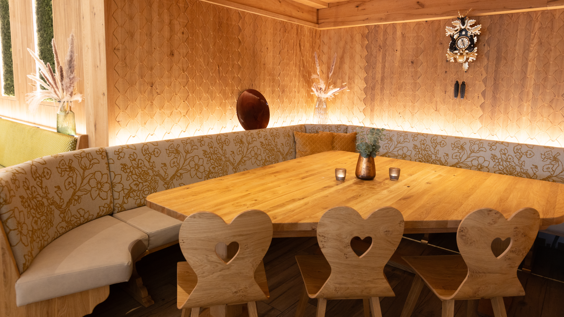 Gemütlicher Essbereich mit einem großen runden Holztisch, von Herzen geformten Holzhockern, einer gepolsterten Bank mit Blumendekor und mit Kerzen sowie Vasen dekoriert, Holzpaneele an der Wand und eine historische Uhr.