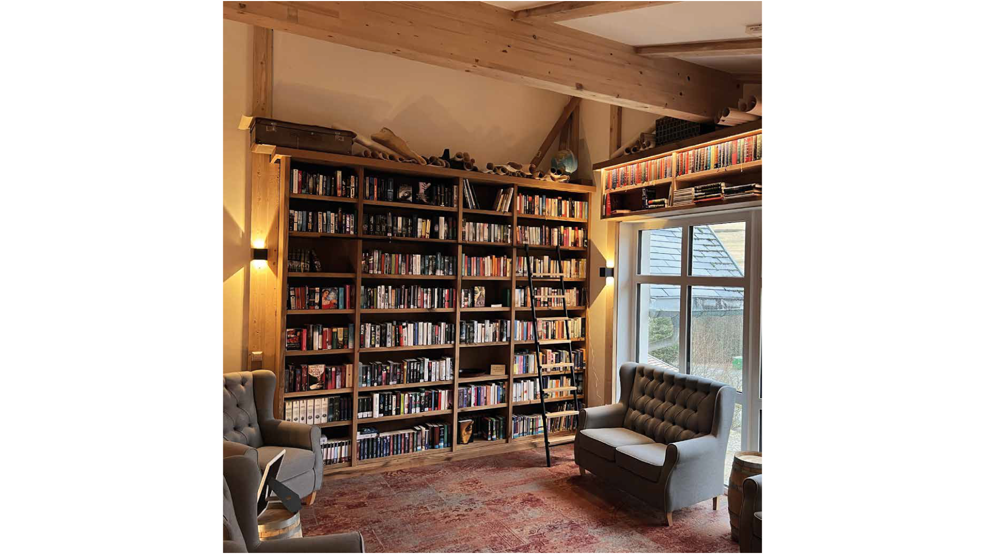 Bücherregal in einem Wohnzimmer, mit einem Treppenschub für Bücher, neben einem großen Fenster, mit zwei Sessel und einem Teppich im Raum.