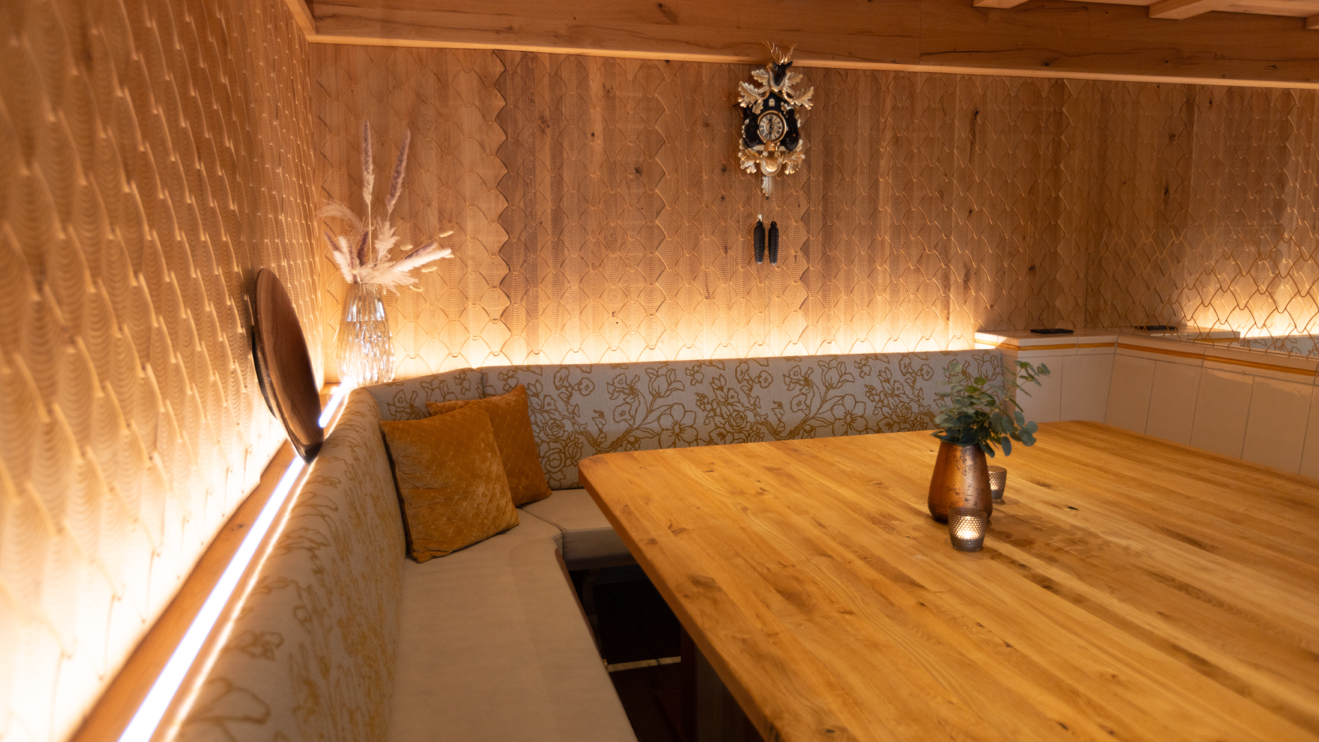 Gemütliches Esszimmer mit Holzdecke, beleuchteten Holzwandpaneelen, einem großen Holztisch mit Vasen und Kerzen, auf einer Seite einer Sitzbank mit Kissen. Es hängt eine antike Uhr an der Wand.
