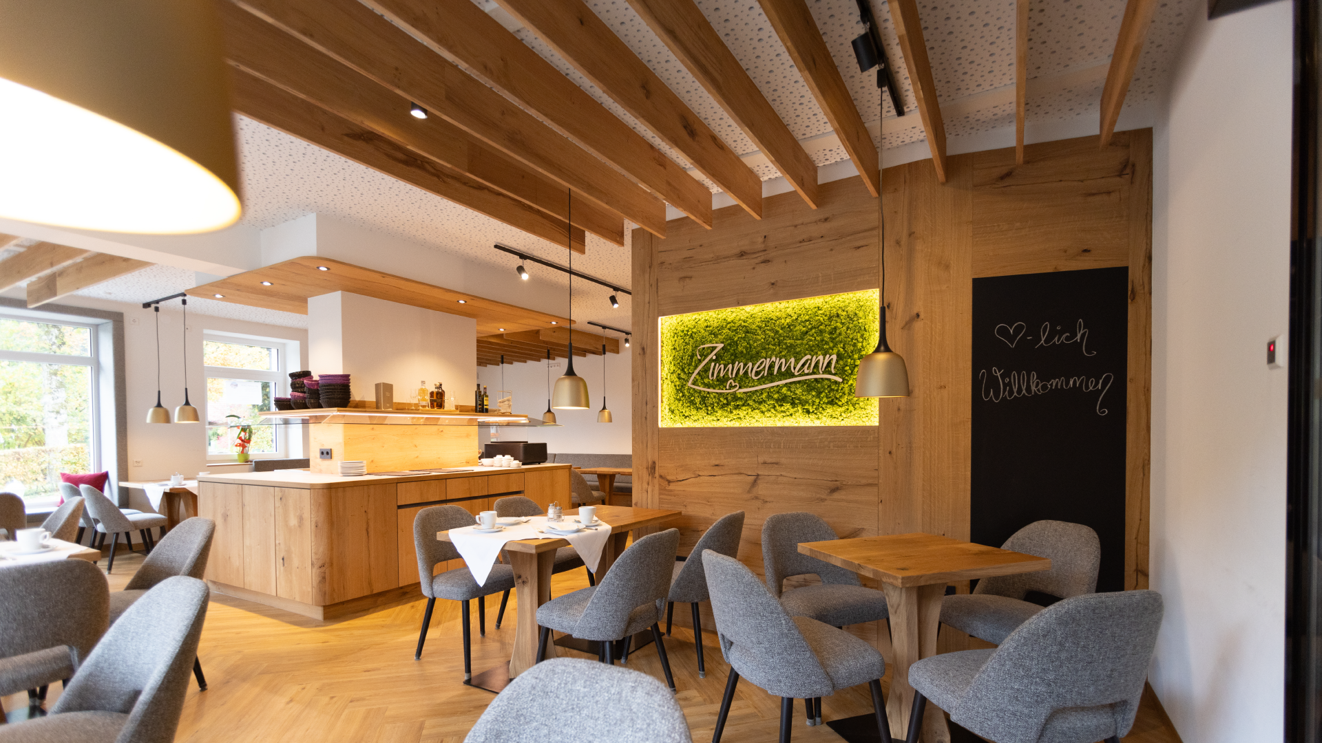 Gemütliches Restaurant mit Holzmöbeln, grauen Sesseln, festlich gedeckten Tischen, einer Theke im Hintergrund, grüner Begrüßungsseifenwand mit Schriftzug Zimmermann, Wandtafel mit Willkommensgruß
