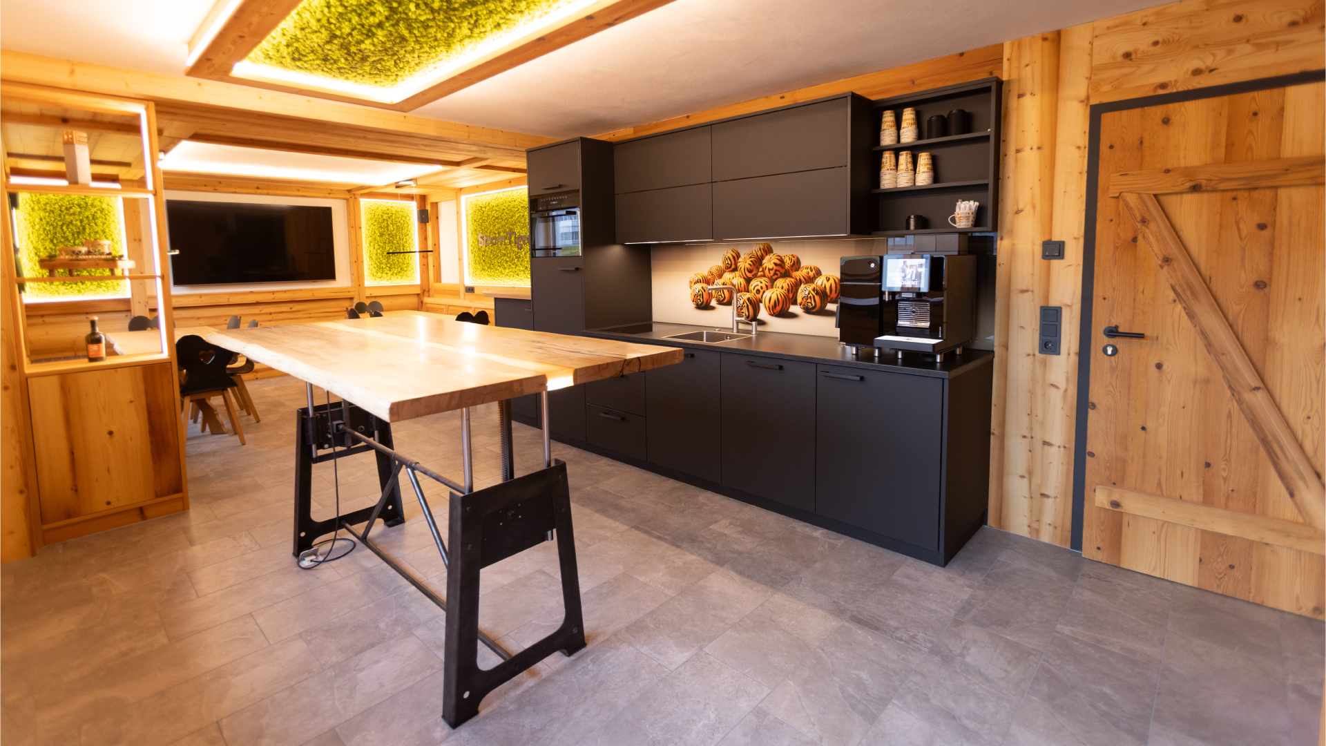 Küche mit schwarzen Schränken, Holztisch, Fernseher und Kaffeemaschine in einem Raum mit Holzverkleidung.
