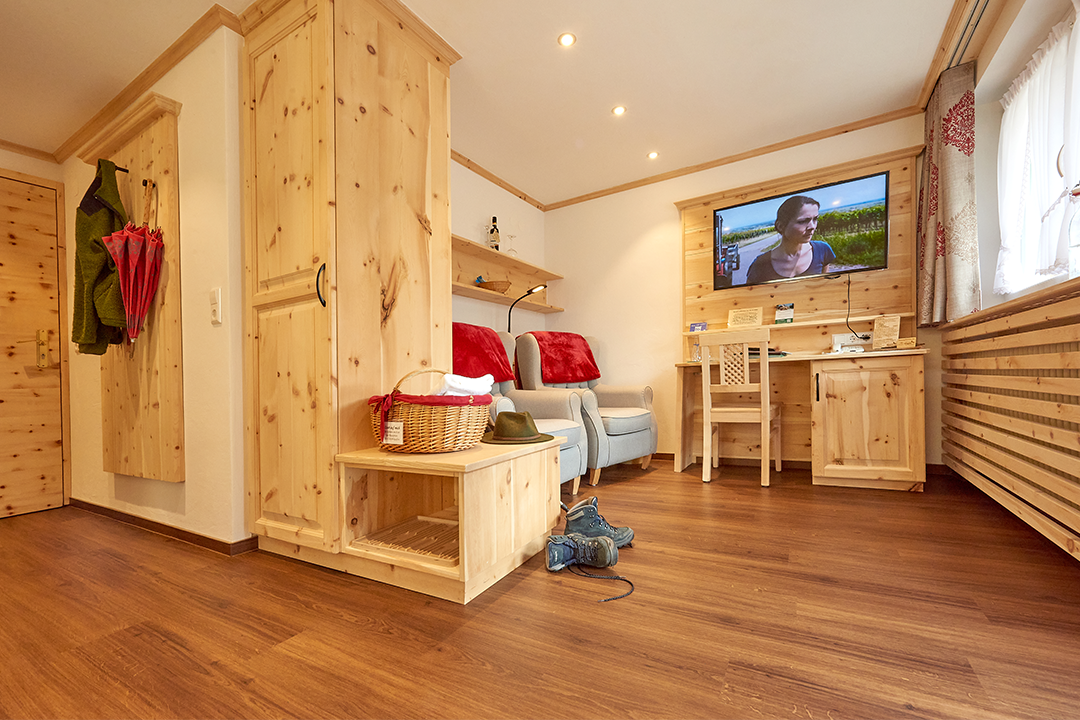 Gemütliches Wohnzimmer mit Holzvertäfelung, zwei bequemen Sesseln, einem Fernseher an der Wand, einem Schreibtisch, Garderobe mit Jacken und Regenschirmen, und Holzboden.